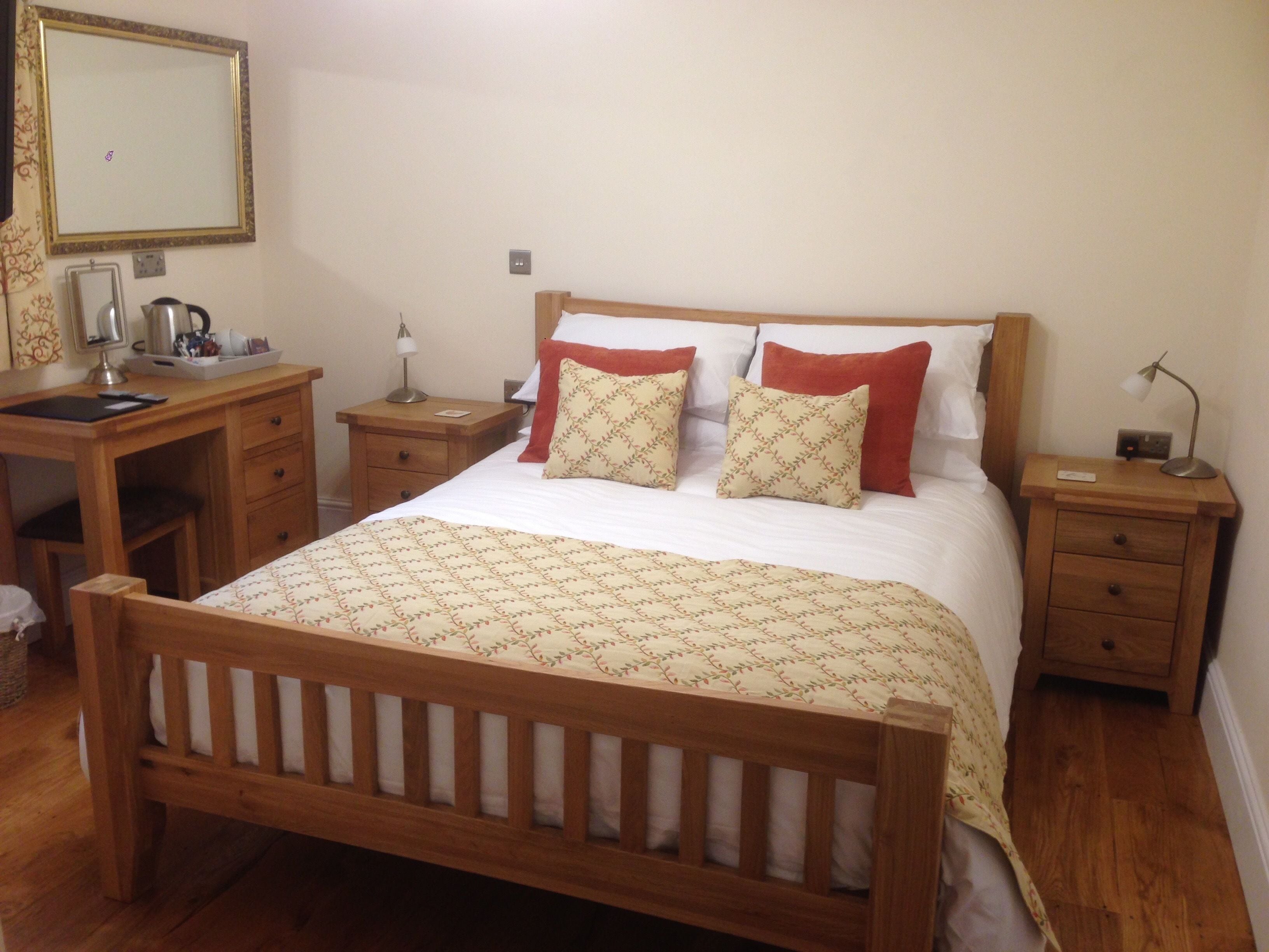 A bedroom with a wooden bed, a desk, and a chair. There is a nightstand with a lamp and a mirror.