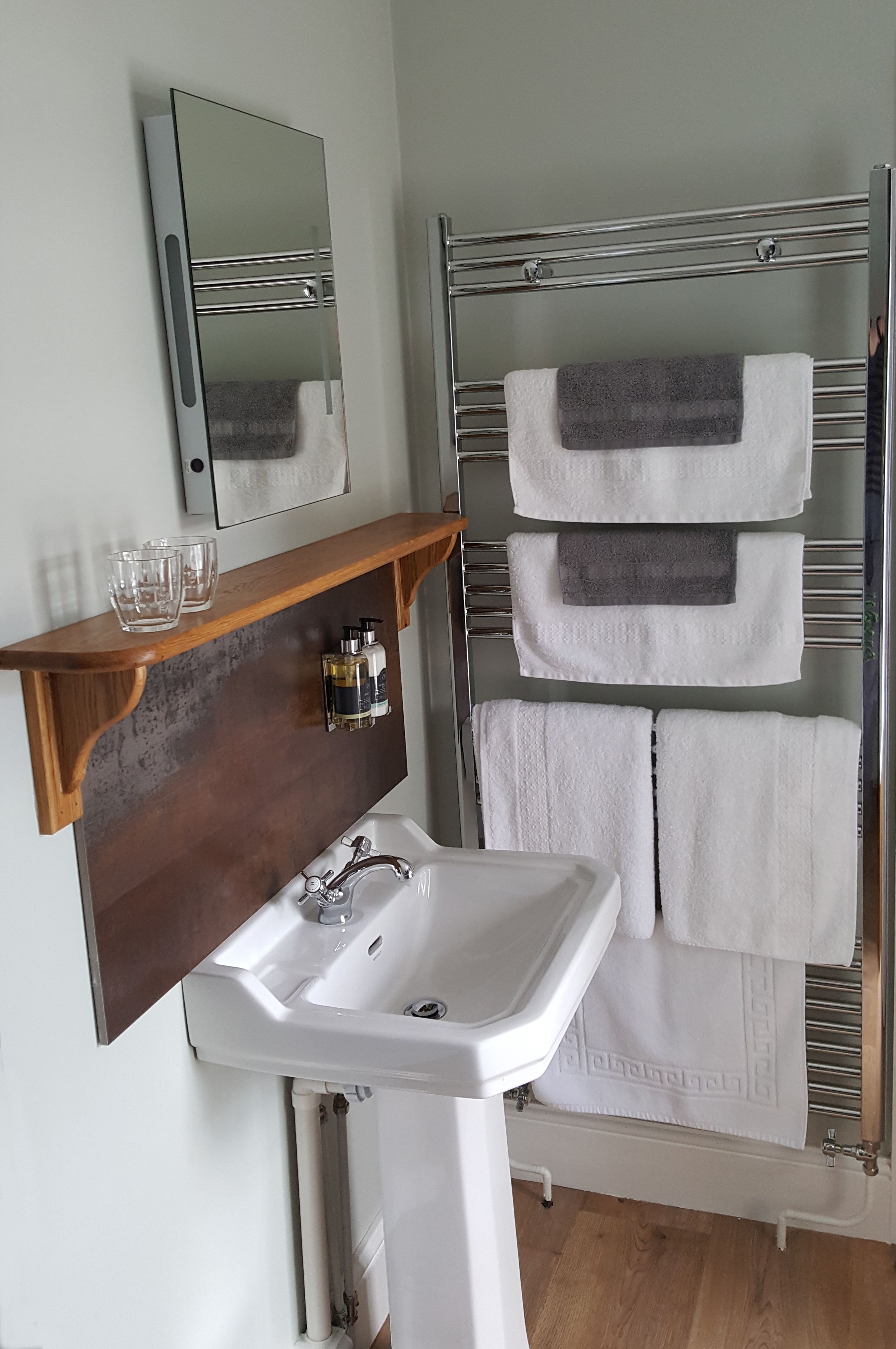 A bathroom with a white sink, a wooden shelf with two glasses, a mirror, and a towel rack with three towels.