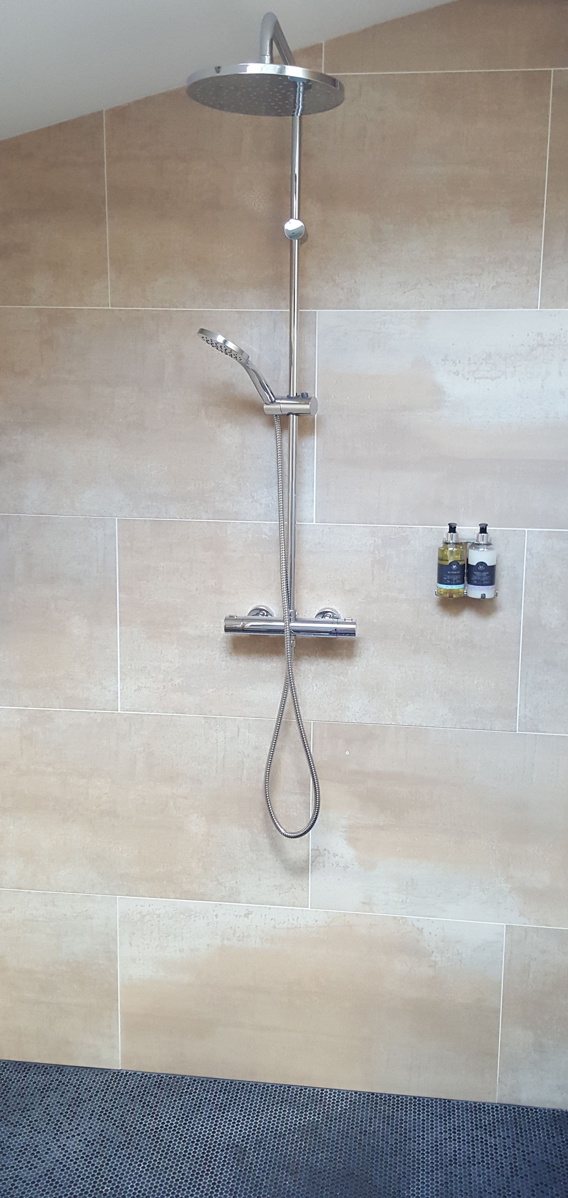 A shower with a handheld showerhead and a rain shower attachment, mounted on a wall with beige tiles.