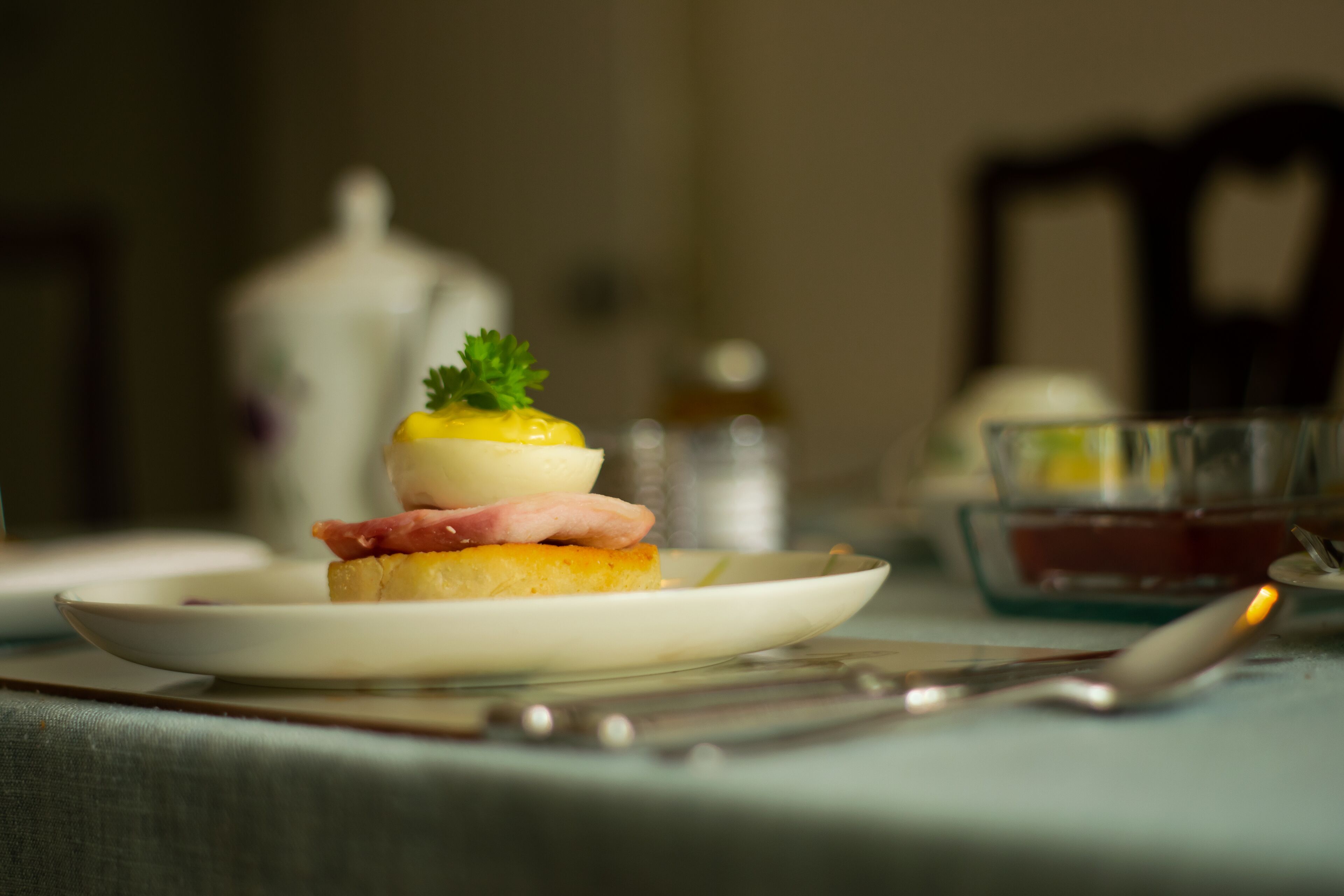 A plate with a sandwich, a boiled egg, and a garnish on a table with a knife and a glass.