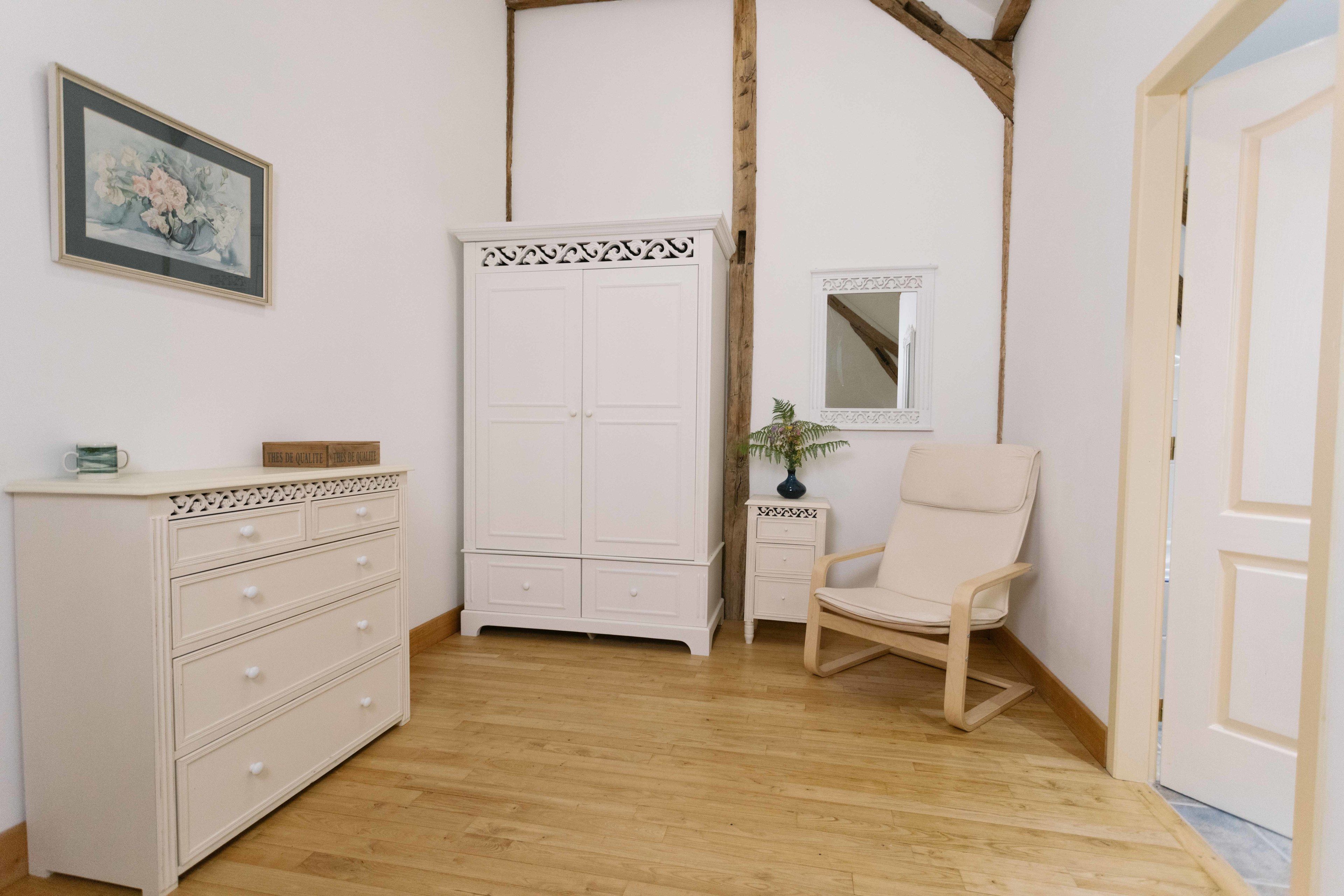 Una habitación con piso de madera, un aparador blanco con cajones, un armario blanco, una silla blanca, una mesita con un florero y un cuadro en la pared.