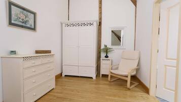 Una habitación con piso de madera, un aparador blanco con cajones, un armario blanco, una silla blanca, una mesita con un florero y un cuadro en la pared.