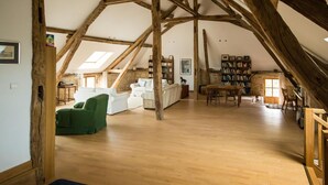 Een ruime zolderkamer met houten balken, een witte bank, een groene fauteuil en een boekenplank.