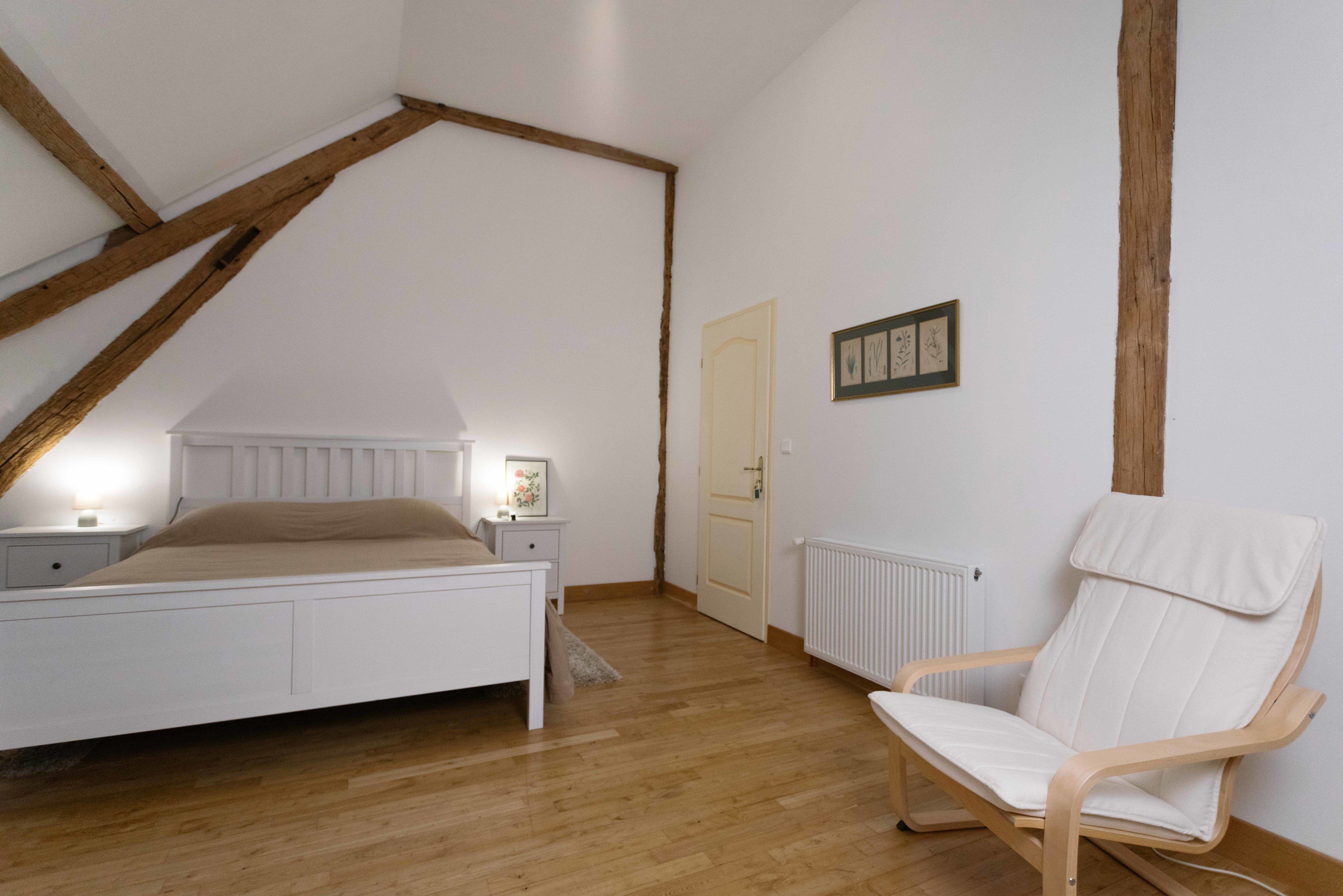 Une chambre avec un plafond en pente, un lit, une table de chevet, une chaise et une porte.