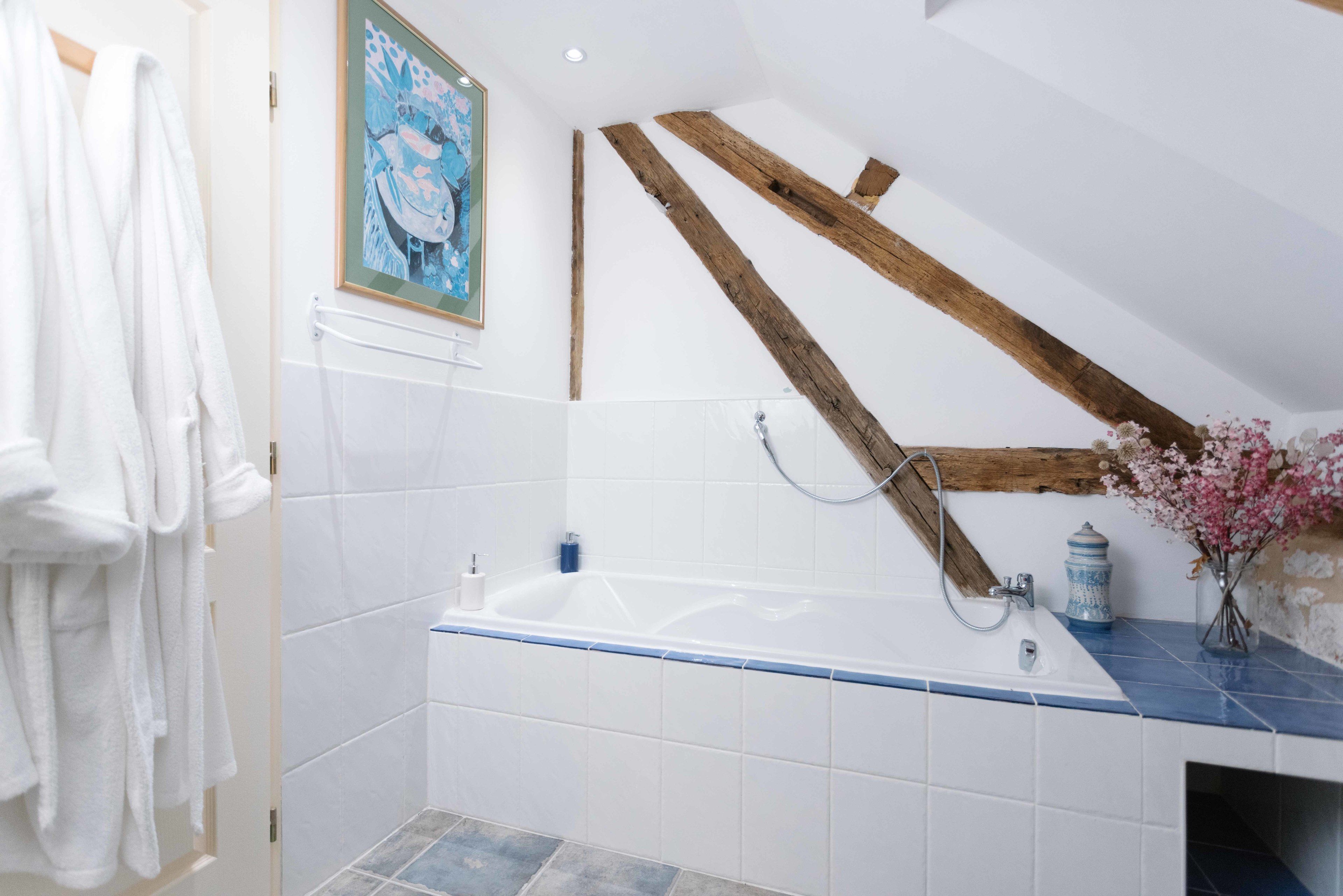 Un bagno con una vasca, piastrelle bianche, travi in legno, un porta asciugamani e un quadro appeso al muro.
