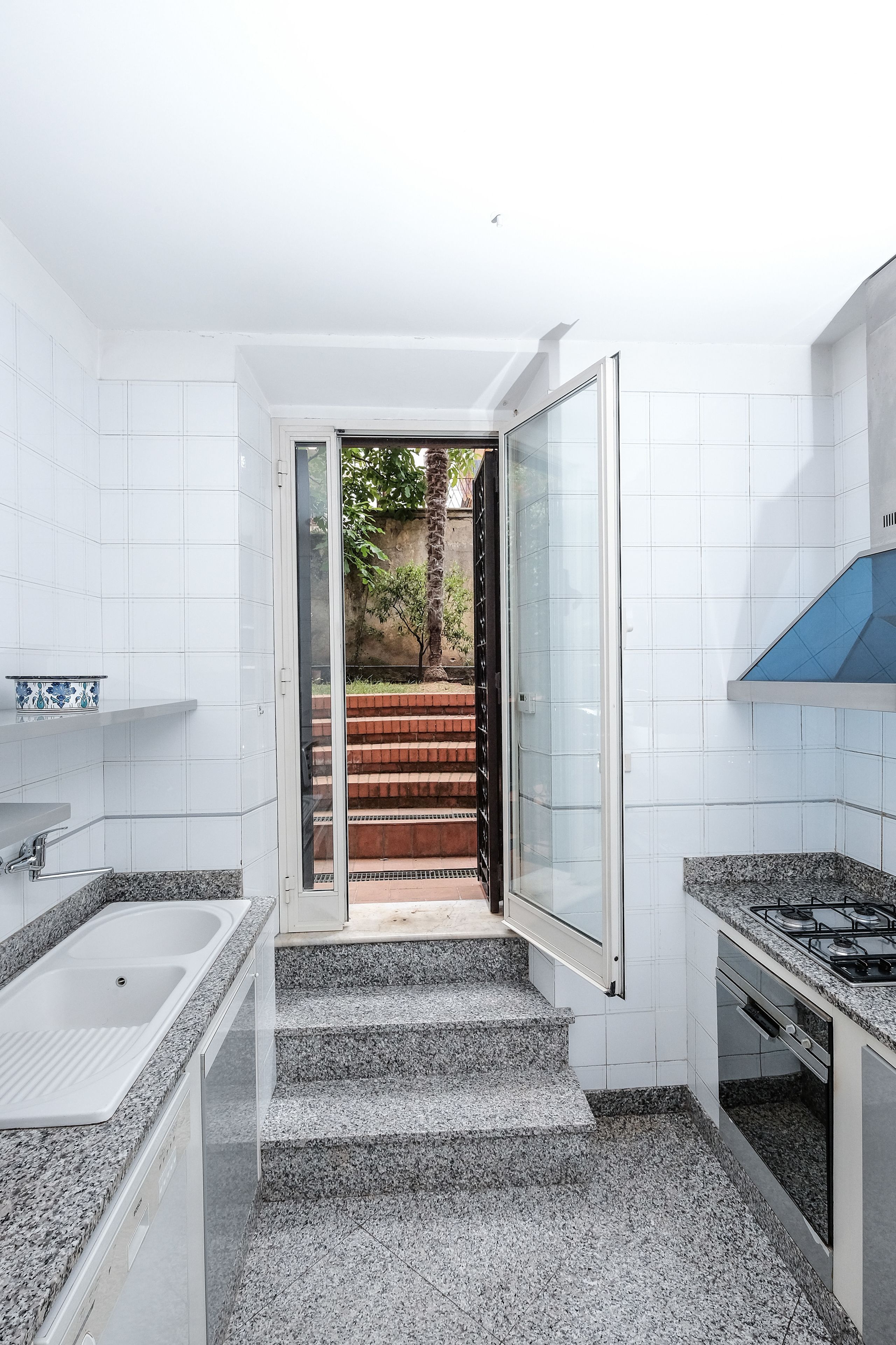 Una cocina moderna con baldosas blancas, encimeras de granito, horno empotrado y puerta de cristal que da a una escalera.