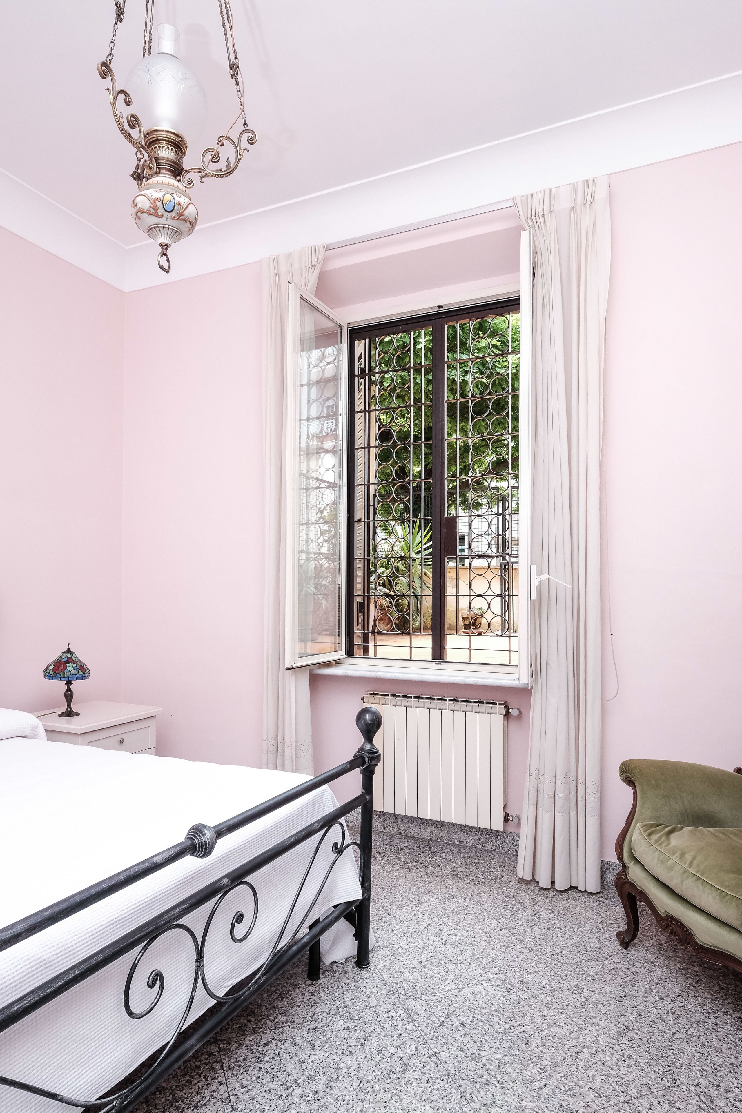 A bedroom with a bed, a radiator, a window with curtains, a green armchair, and a chandelier.