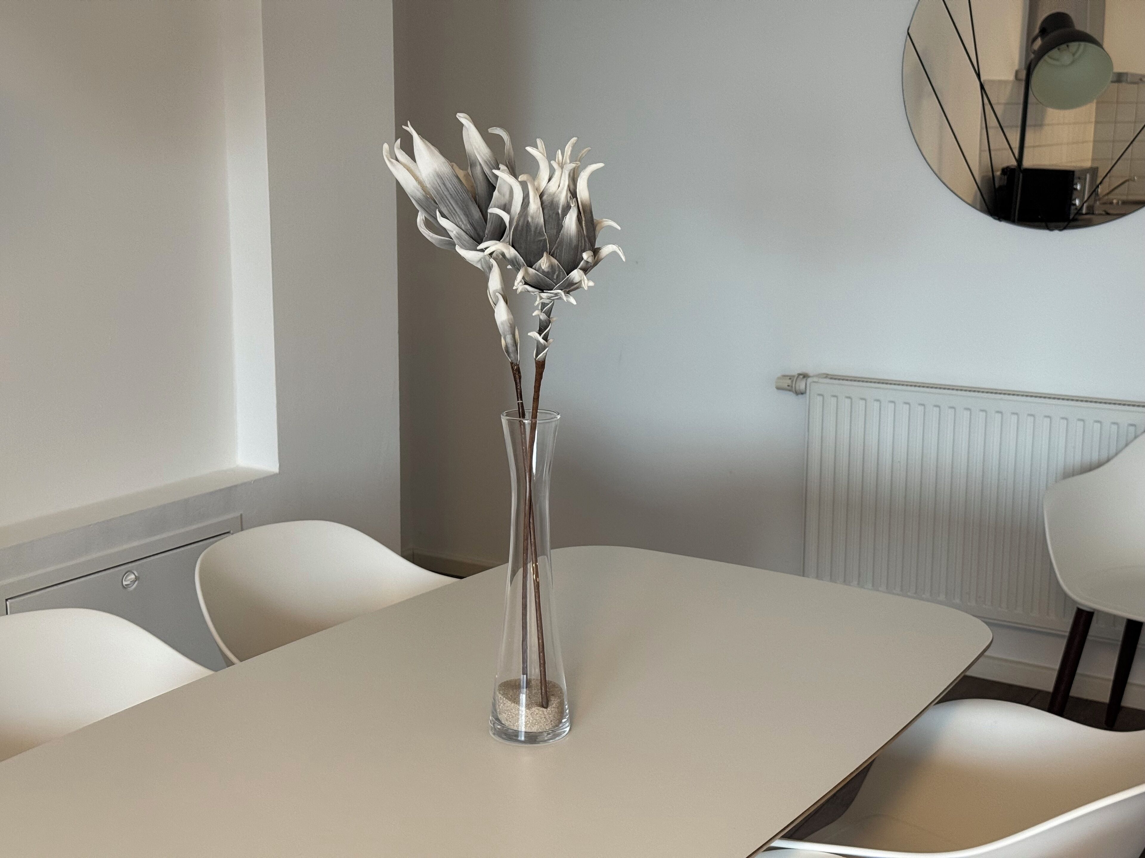 A modern dining area with a white table, clear vase with flowers, and a round mirror on the wall.