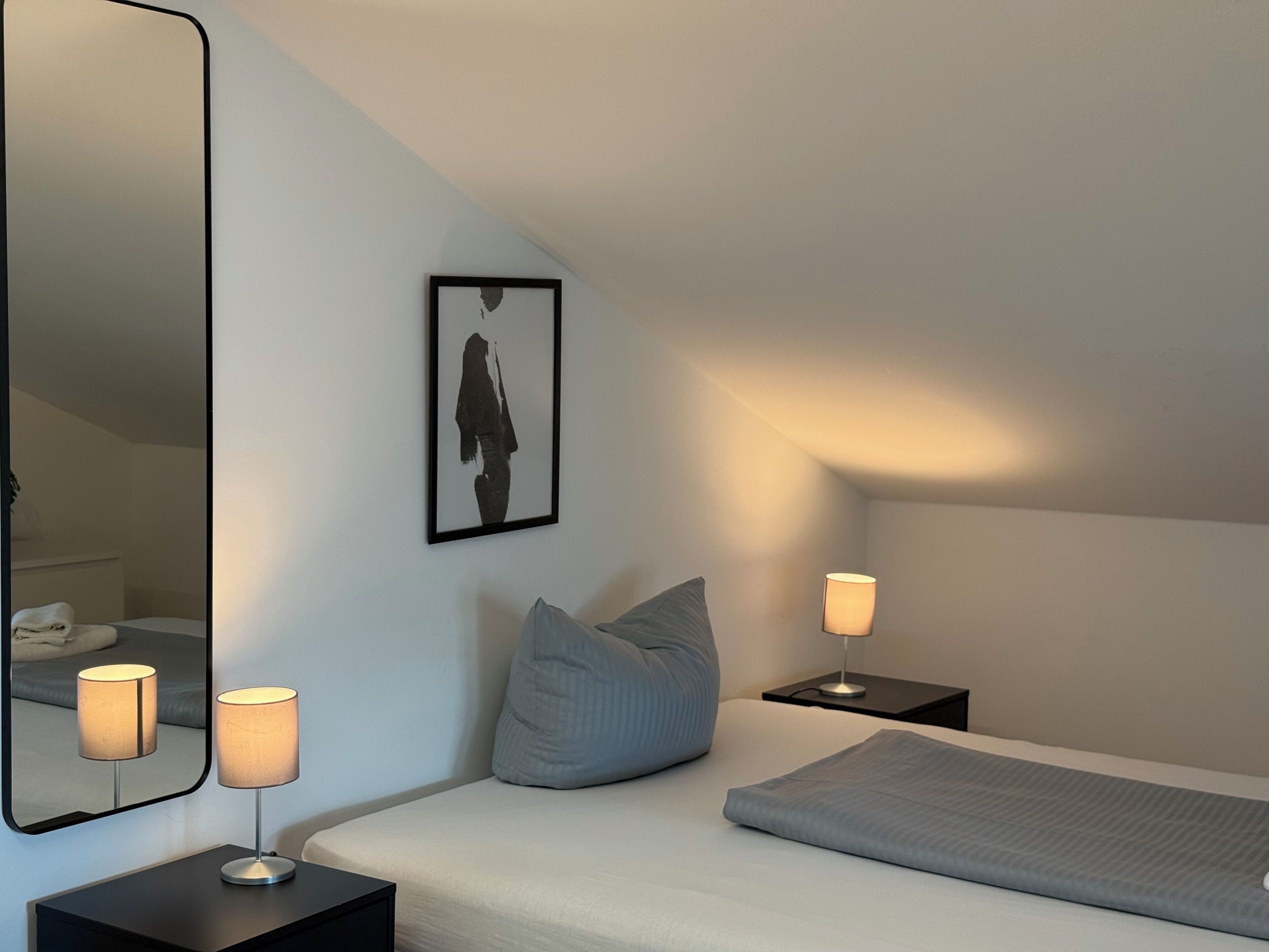 Ein modernes Schlafzimmer mit einem großen Bett, einem Nachttisch mit Lampe, einem weiteren Nachttisch mit Lampe, einem gerahmten Bild an der Wand und einem Spiegel.