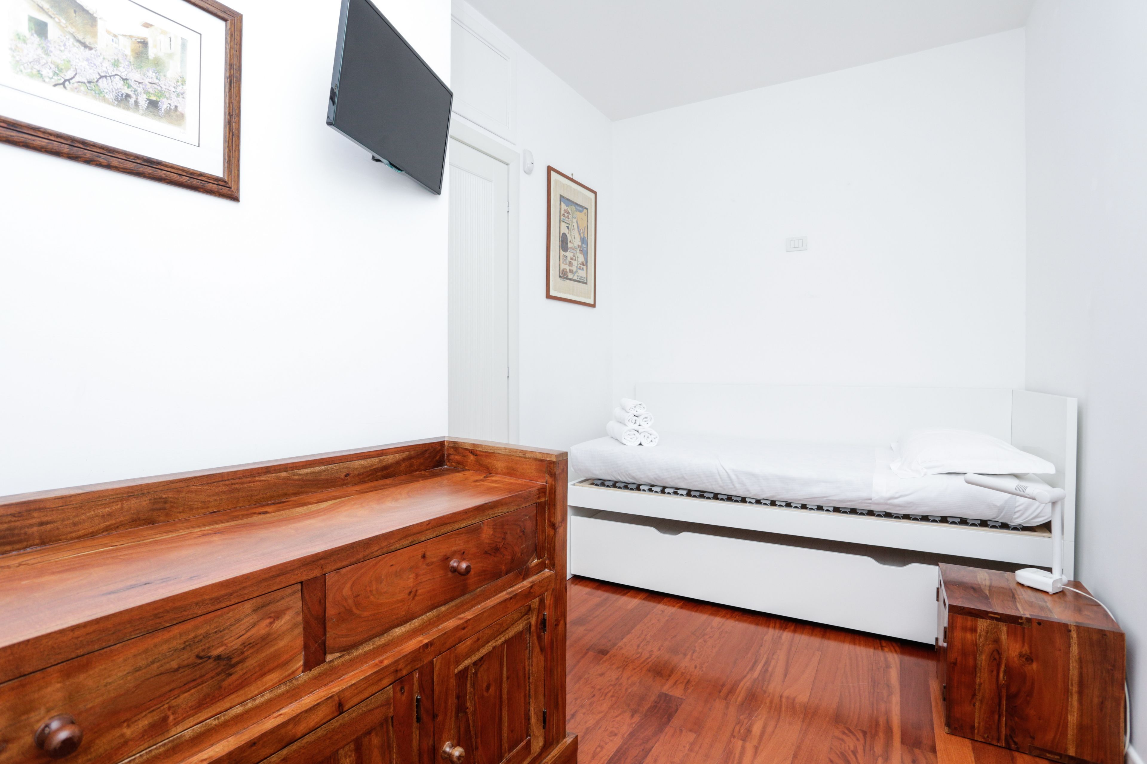 Ein Schlafzimmer mit einem Holzkasten, einem Bett, einer weißen Wand und einem an der Wand befestigten Fernseher.