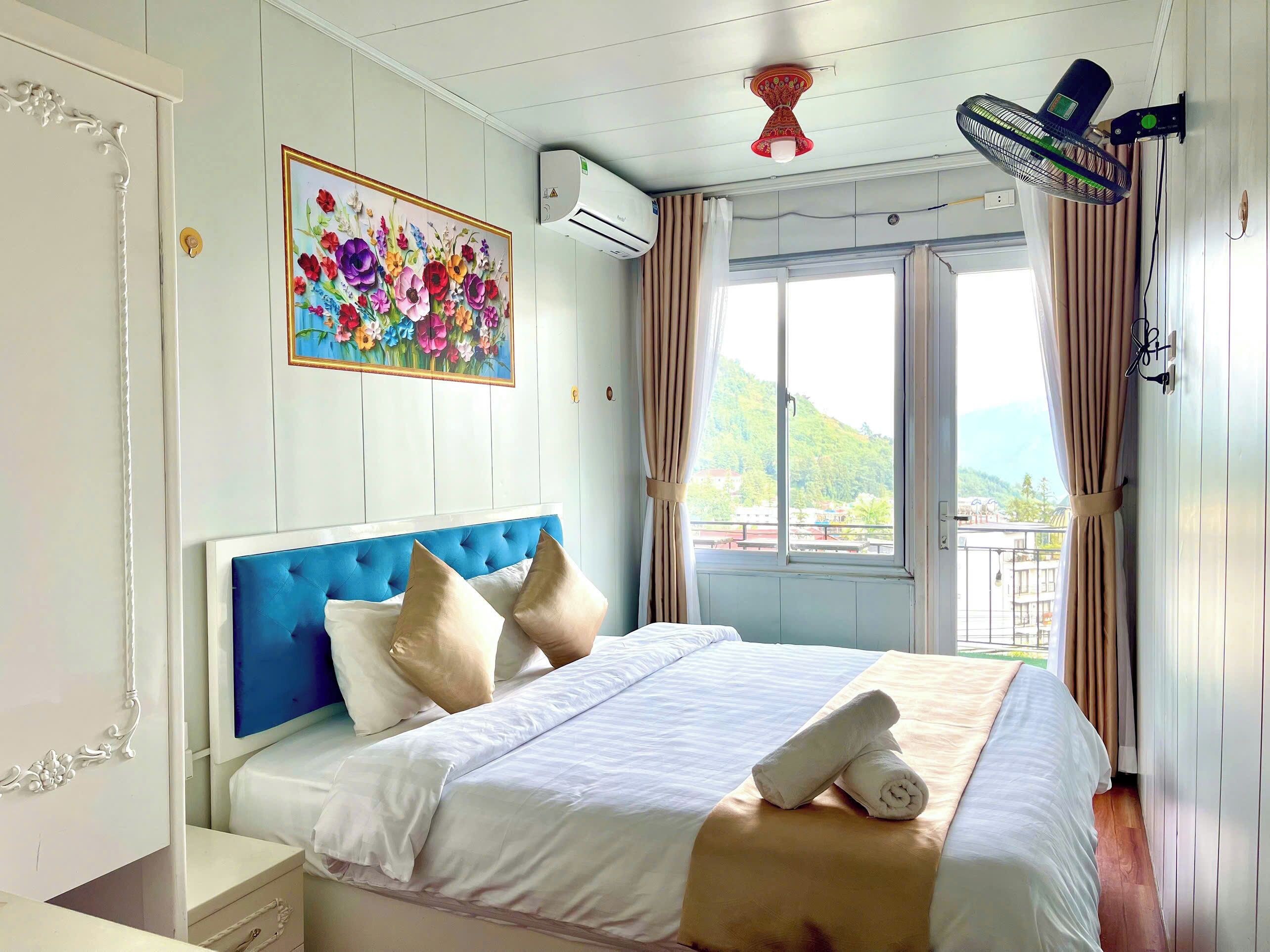 Quarto de hotel com cama, pintura, ar condicionado, ventoinha no teto e janela com cortinas.