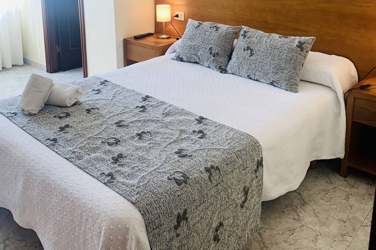 Een net opgemaakt bed met een bedekking met patroon, een houten hoofdbord en twee aan de muur bevestigde bloemstillevens.