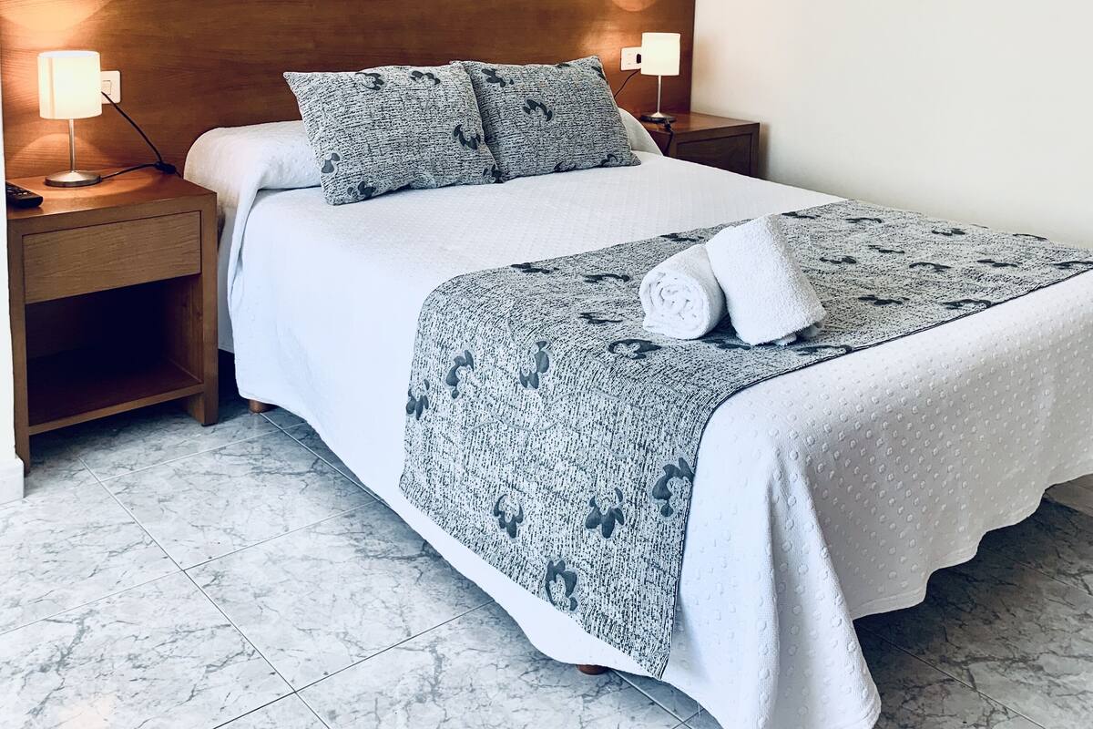 Een net opgemaakt bed met een gebonden deken, twee kussens en een opgevouwen handdoek. Twee nachtlampjes, een houten nachtkastje en een marmeren vloer.