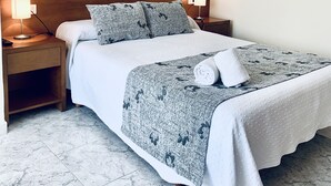 Ein ordentlich bezogenes Bett mit einer gemusterten Decke, zwei Kissen und einem gefalteten Handtuch. Zwei Nachttischlampen, ein Nachttisch aus Holz und ein Boden mit Marmorfliesen sind ebenfalls zu sehen.
