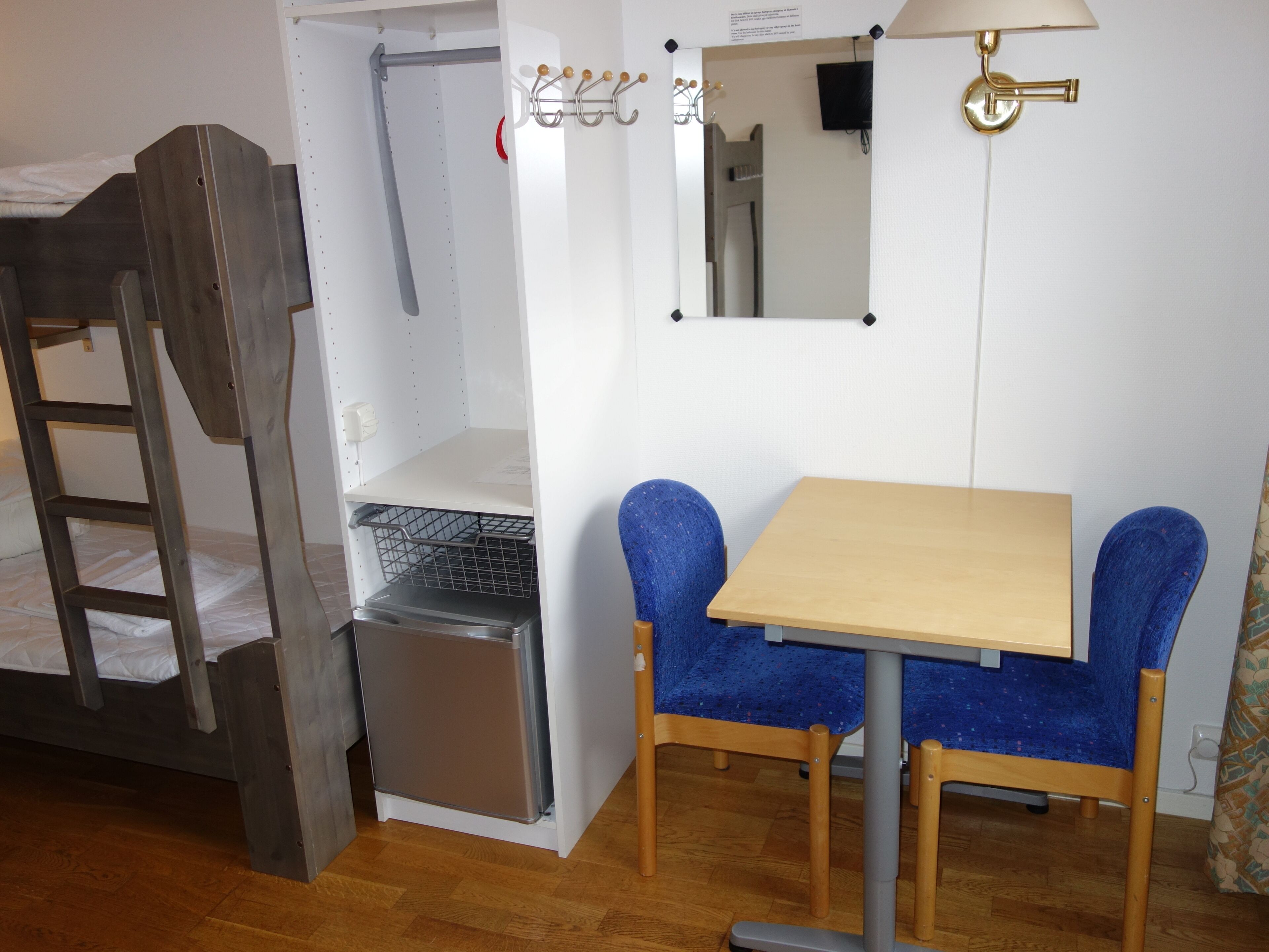 A compact room with a bunk bed, a small refrigerator, a table with two chairs, and a wall-mounted lamp.