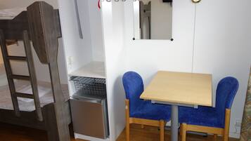 A compact room with a bunk bed, a small refrigerator, a table with two chairs, and a wall-mounted lamp.