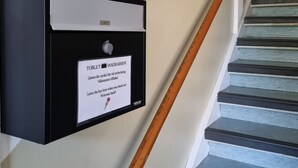 A black intercom box with a plant on top, a sign with instructions, and a wooden handrail beside a staircase. - Torget Vandrarhem - Hostel (Kavlinge)