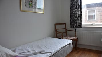 A small, single-bed room with a wooden chair, a radiator, and a window with curtains.