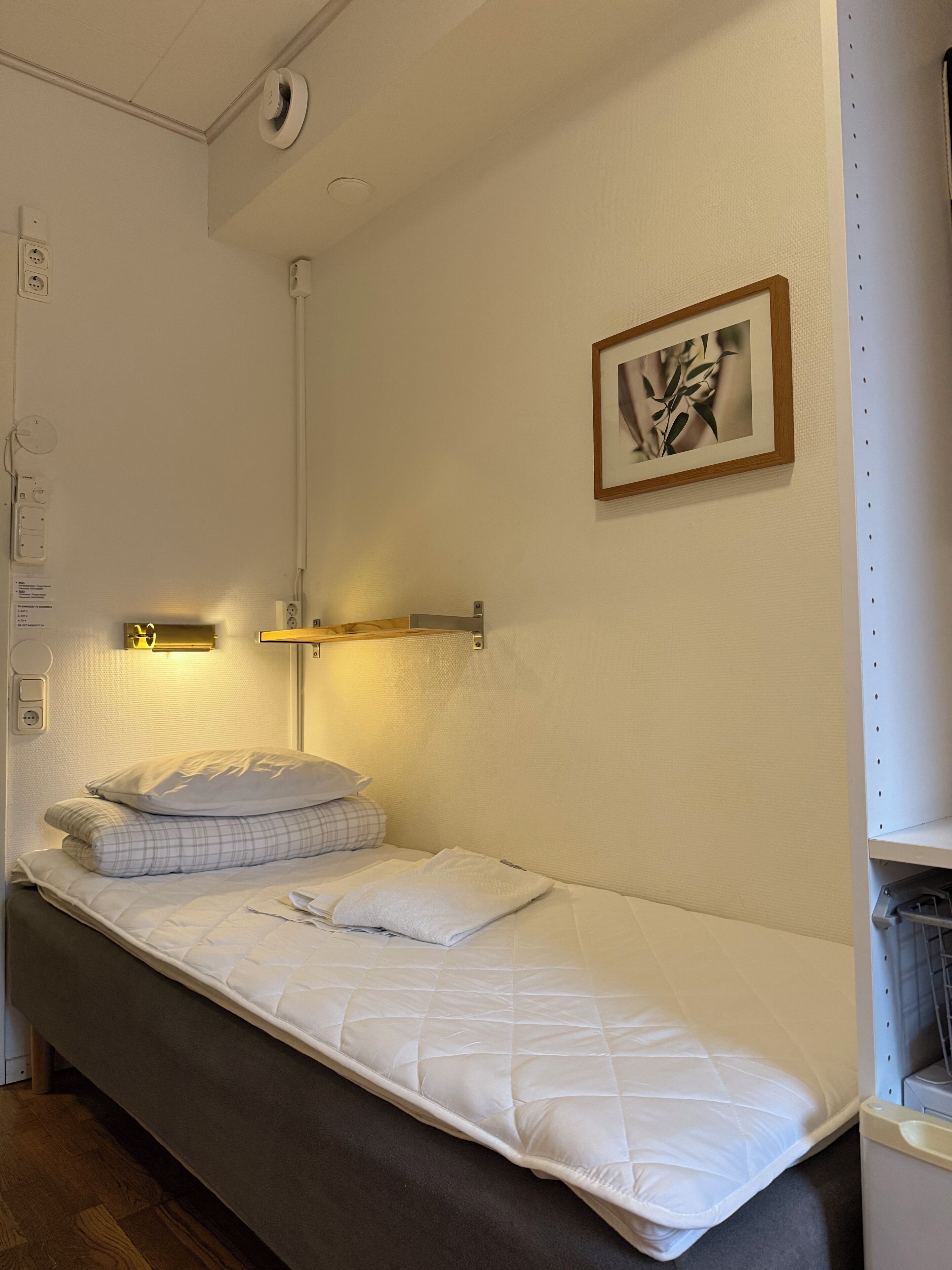 Una cama individual con colchón acolchado, una mesita de noche con lámpara, y un cuadro en la pared.