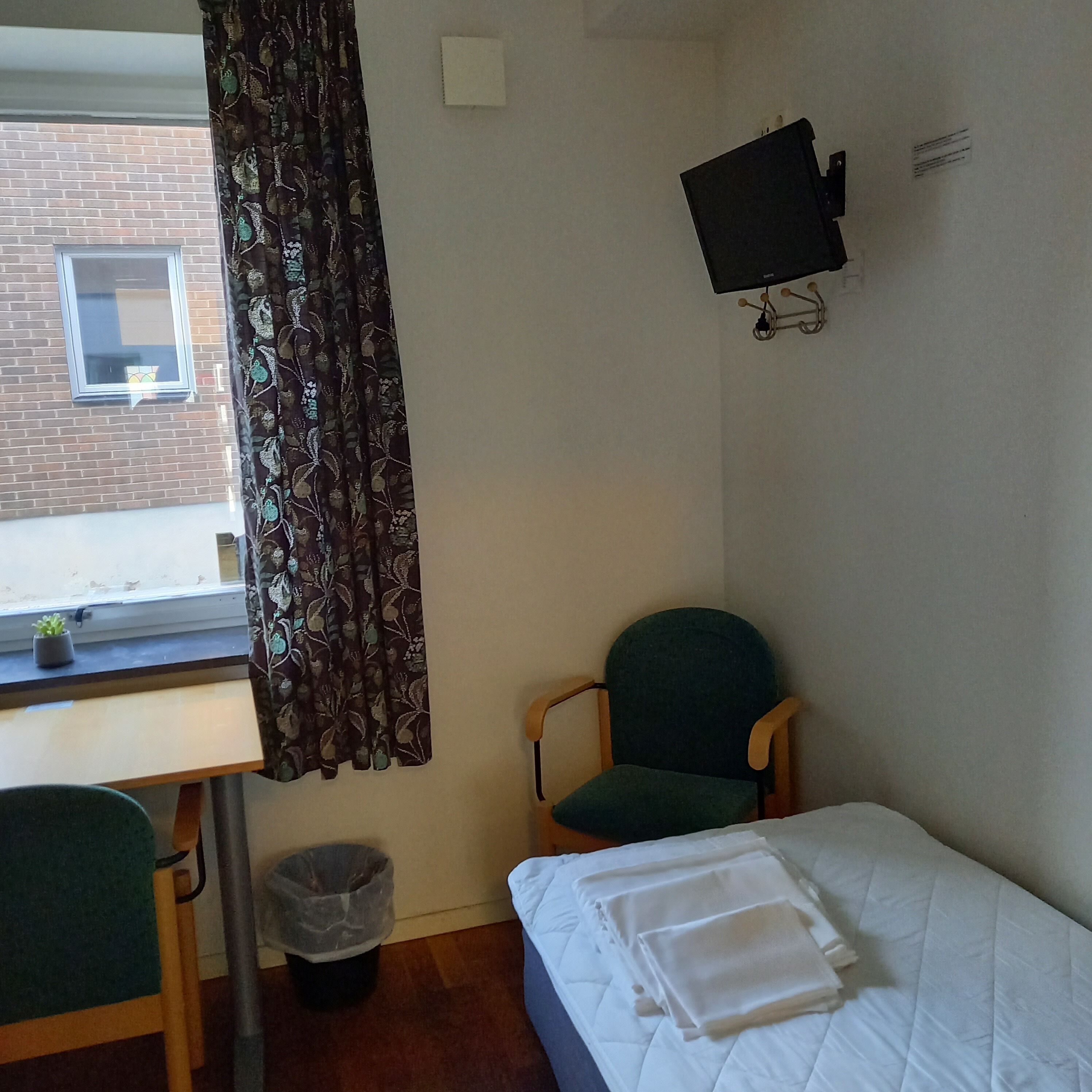 A room with a bed, a desk, a chair, a TV mounted on the wall, and a window with curtains.