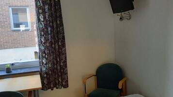 A room with a bed, a desk, a chair, a TV mounted on the wall, and a window with curtains.