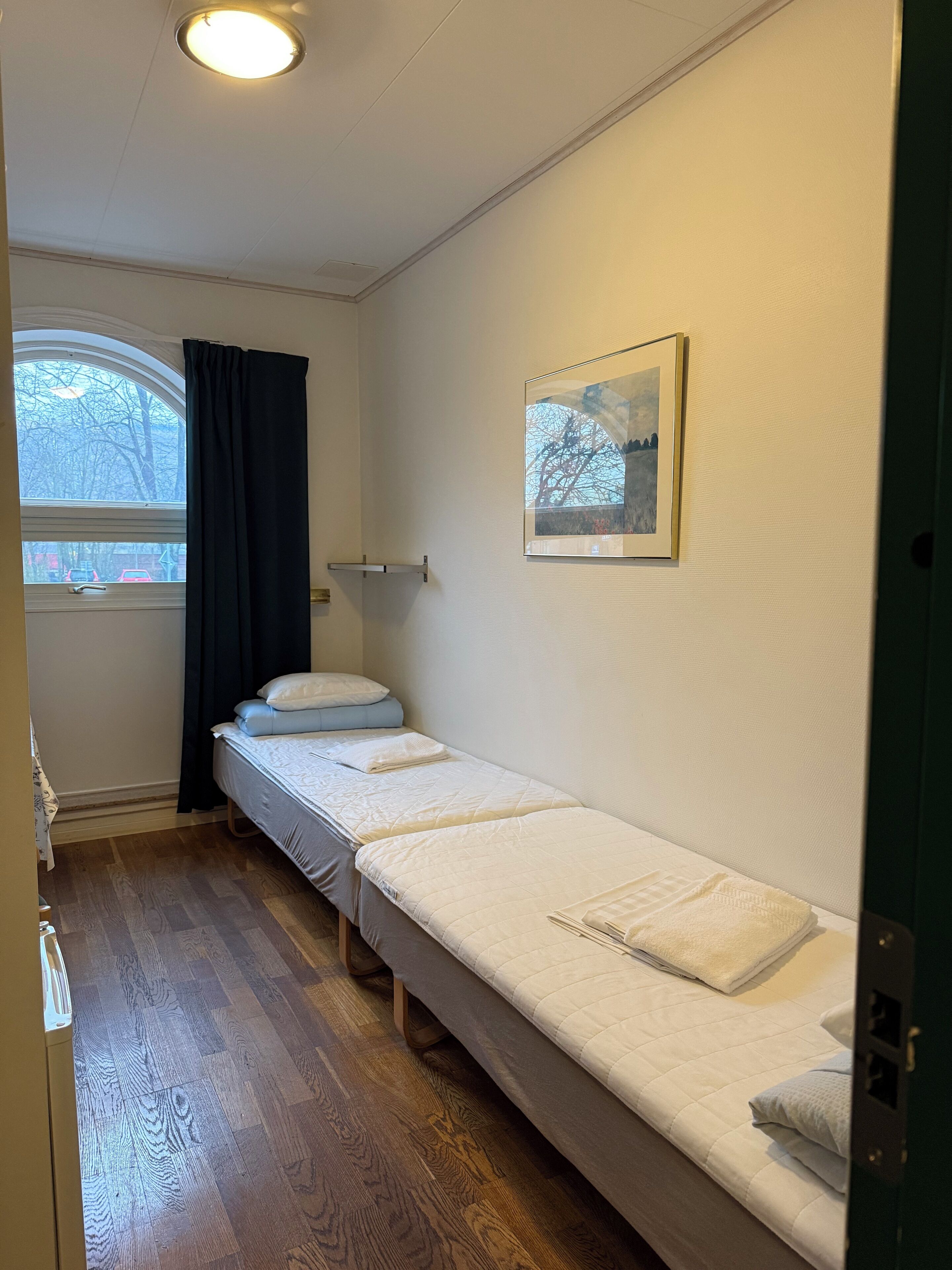 A small, clean room with two single beds, a window with curtains, a framed picture on the wall, and a wooden floor.