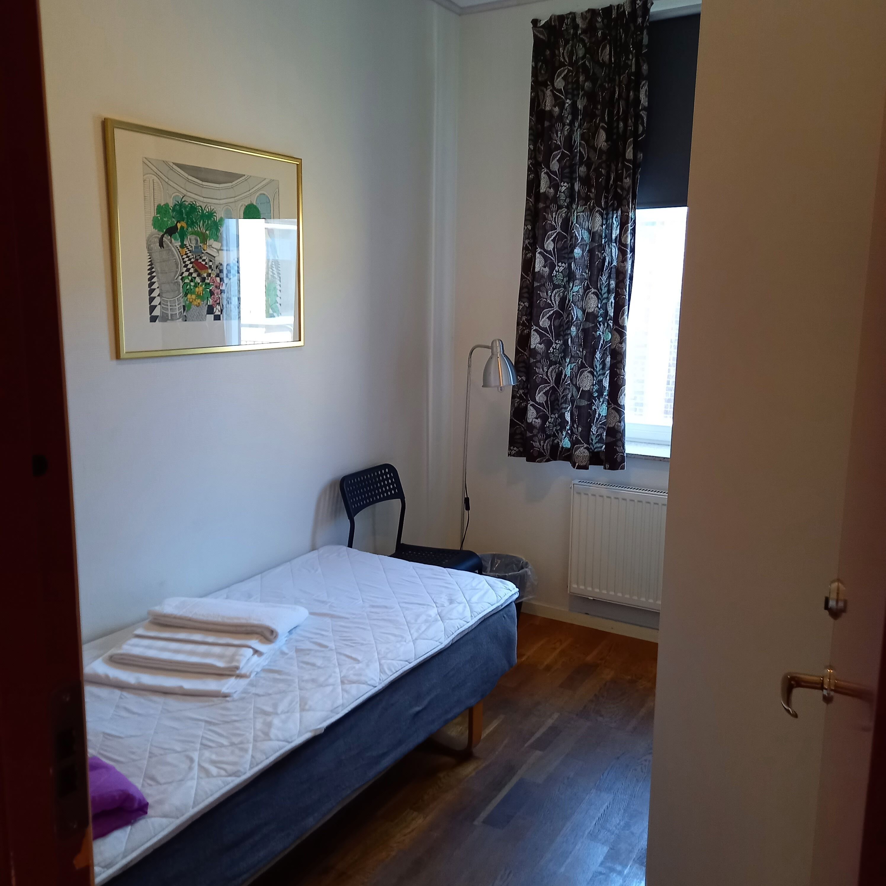 A small, single-bed room with a white mattress, a chair, a radiator, and a window with curtains.