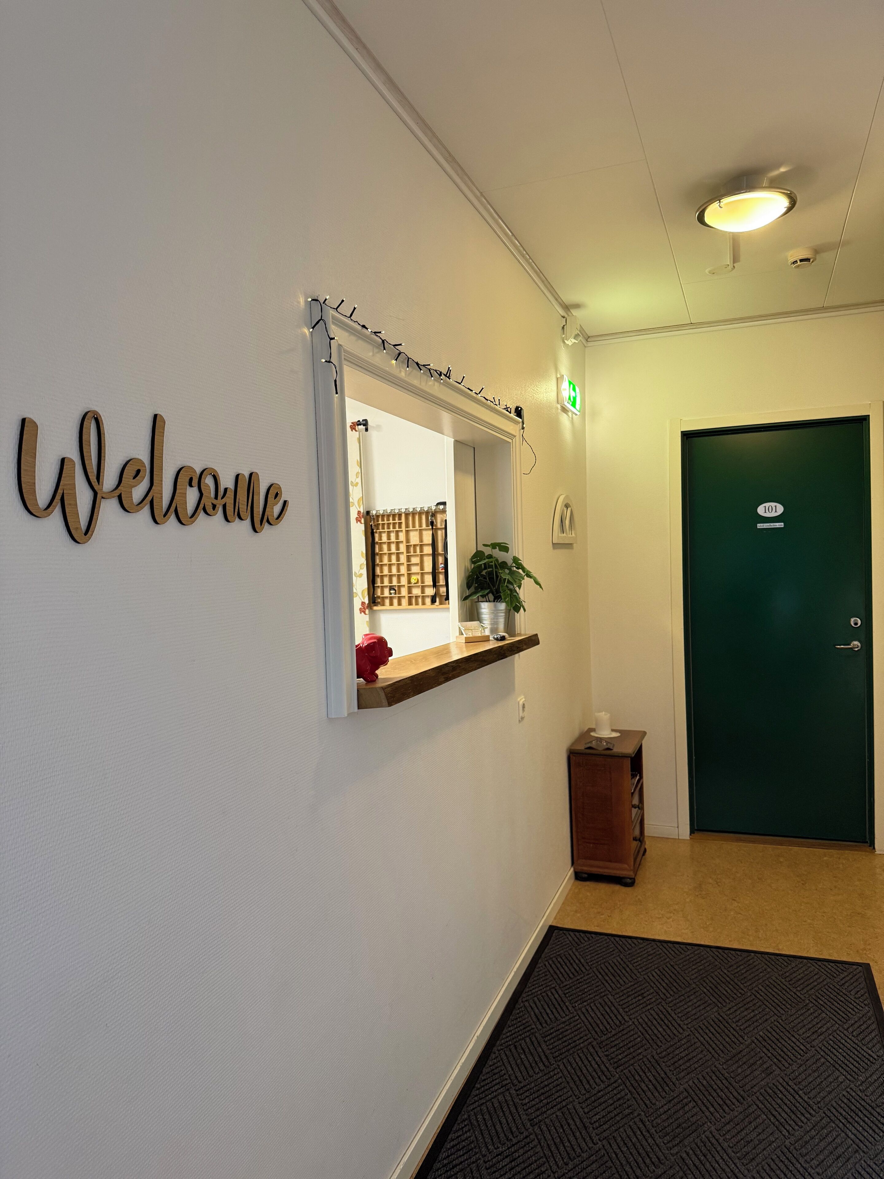 Una zona de recepción con un letrero que dice 'Welcome', un espejo con repisa, una puerta verde y una planta pequeña.