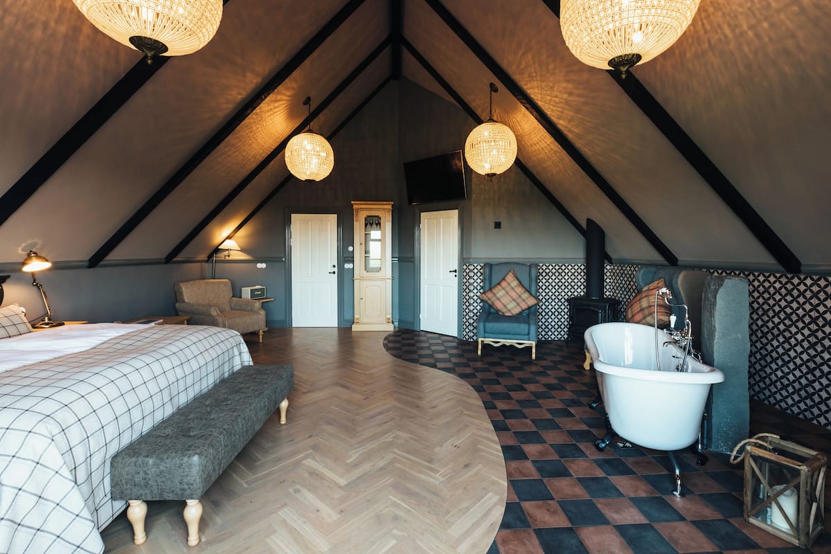 Un dormitorio amplio en el ático con una cama grande, un banco, una bañera de patas de león y una chimenea.