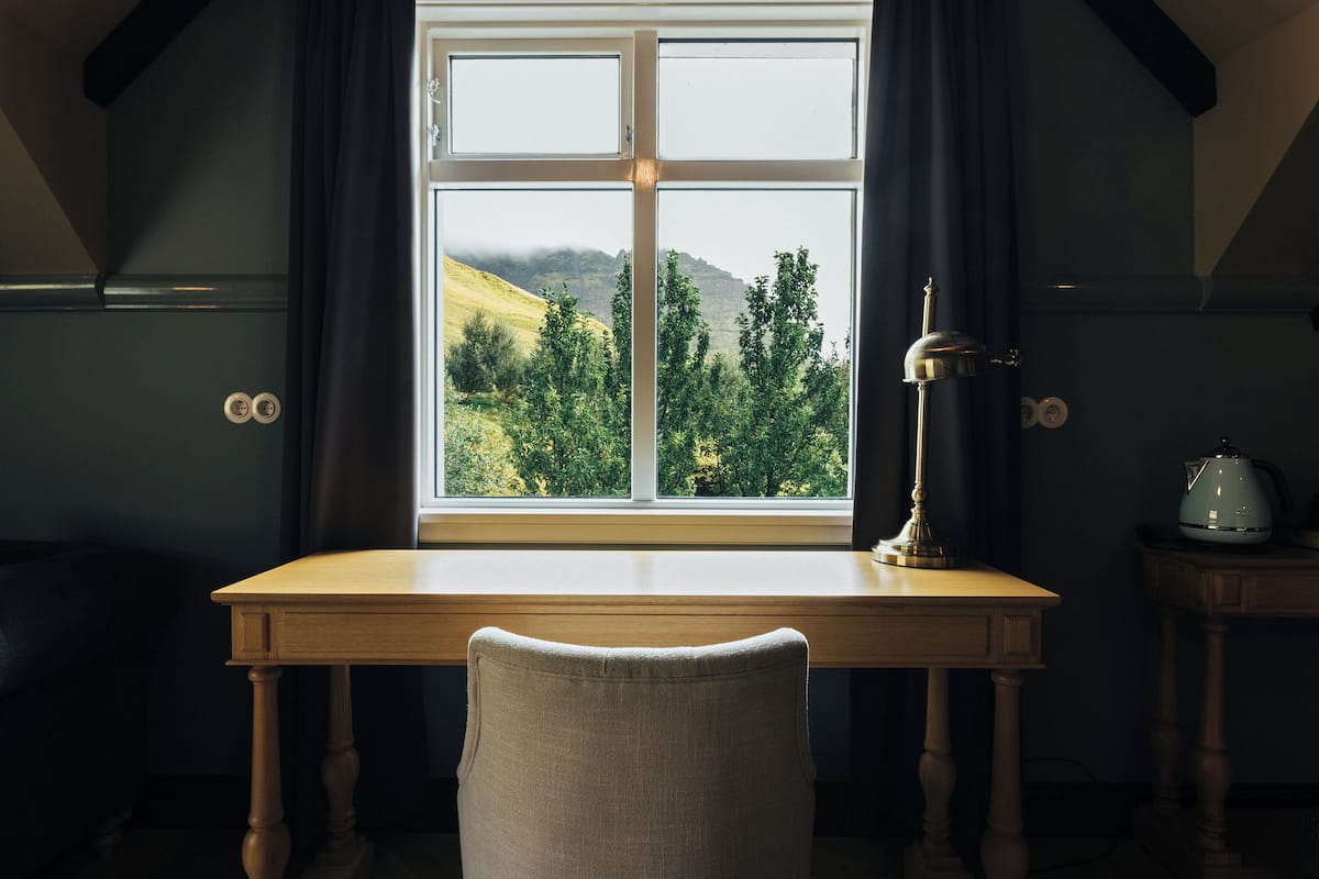 Una habitación con un escritorio de madera, una silla, un ventanal con vistas a un paisaje verde, y una lámpara sobre el escritorio.