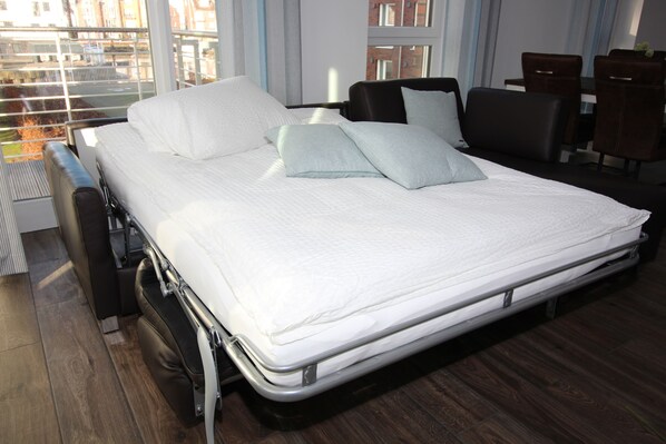 Ein ordentlich bezogenes Bett mit Matratze, Kissen und Kopfteil in einem Hotelzimmer mit Blick auf Gebäude durch große Fenster.