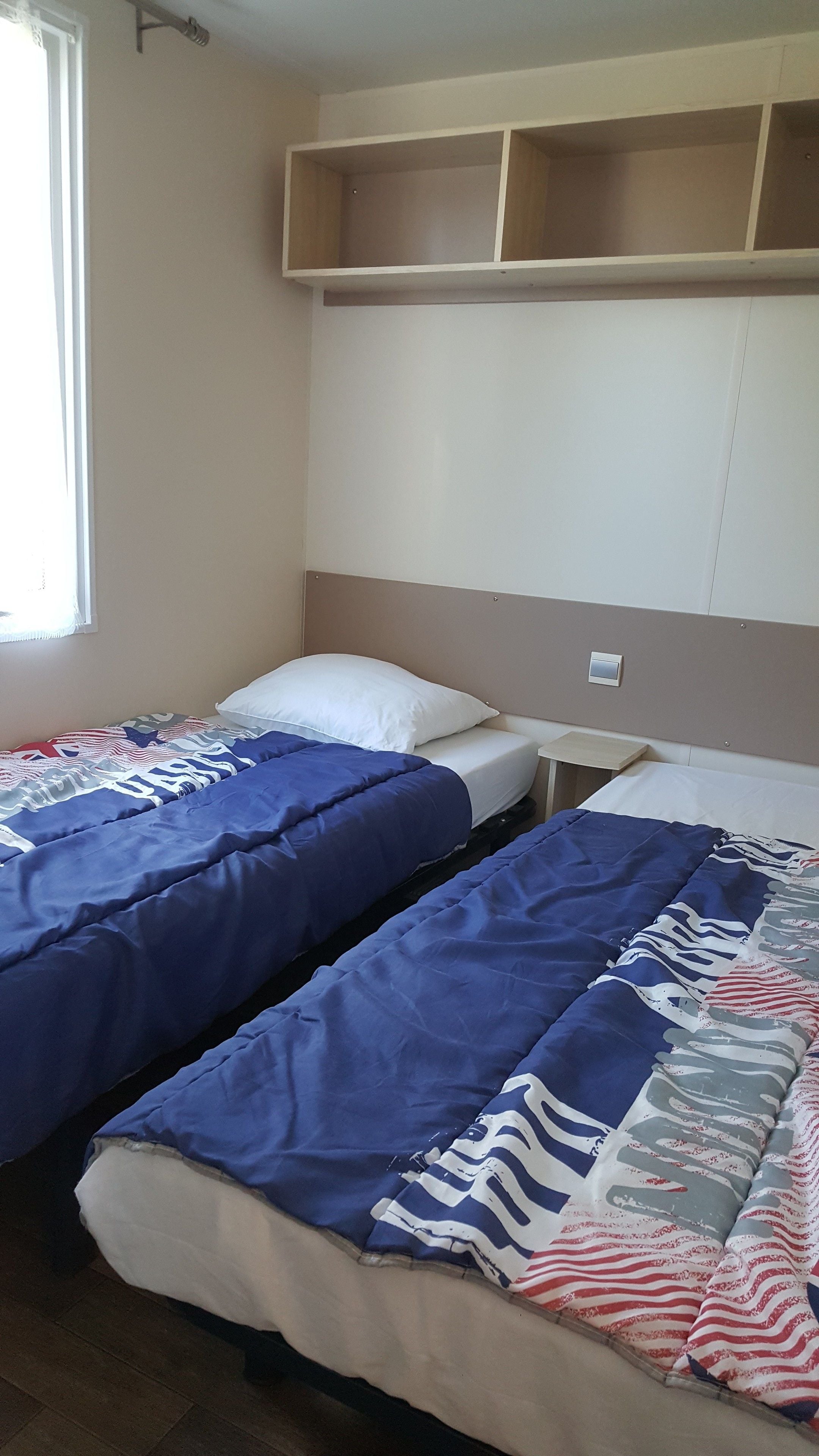 Une chambre avec deux lits, chacun étant équipé de draps bleus et d’un oreiller blanc. Il y a une fenêtre sur le côté gauche et une étagère au-dessus des lits.