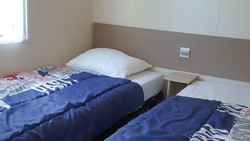 Une chambre avec deux lits, chacun étant équipé de draps bleus et d’un oreiller blanc. Il y a une fenêtre sur le côté gauche et une étagère au-dessus des lits.