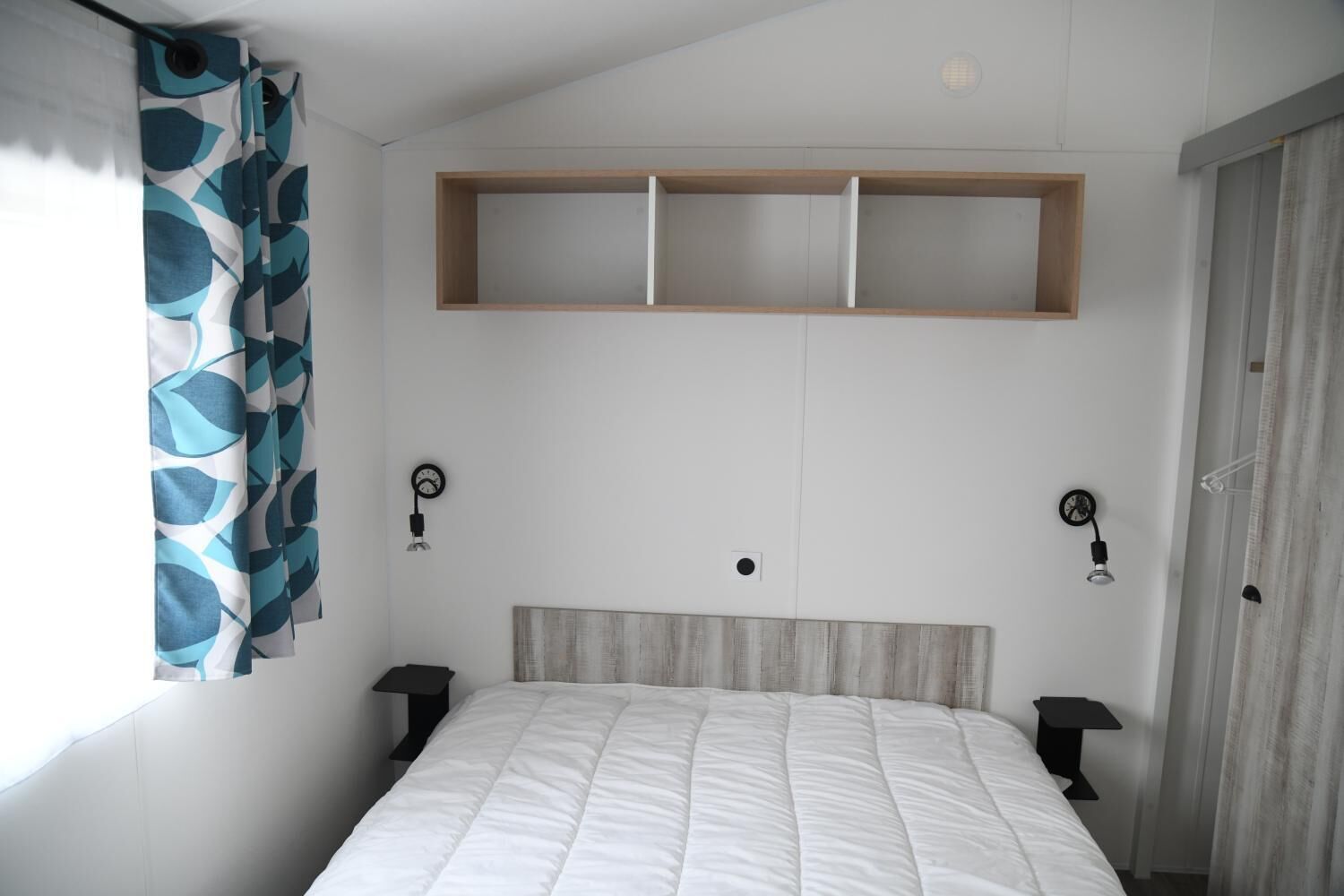Een slaapkamer met een bed, hoofdeinde, twee nachtkastjes, een wandplank en een raam met gordijnen.