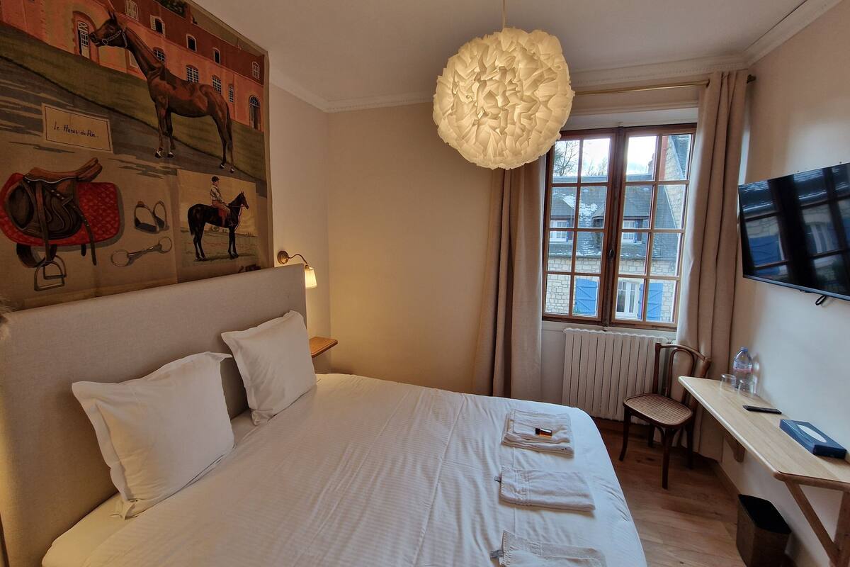 A hotel room with a large bed, a desk, a chair, a TV, and a window with a view of buildings.