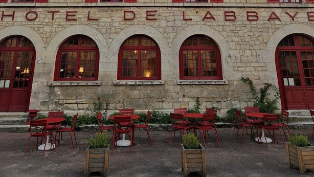 A stone building with 'HOTEL DE L'ABBAYE' signage, arched windows, red shutters, and outdoor seating. - Hotel De L'Abbaye (Longpont)