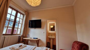 A hotel room with a bed, a TV, a mirror, a red chair, and a window with curtains.