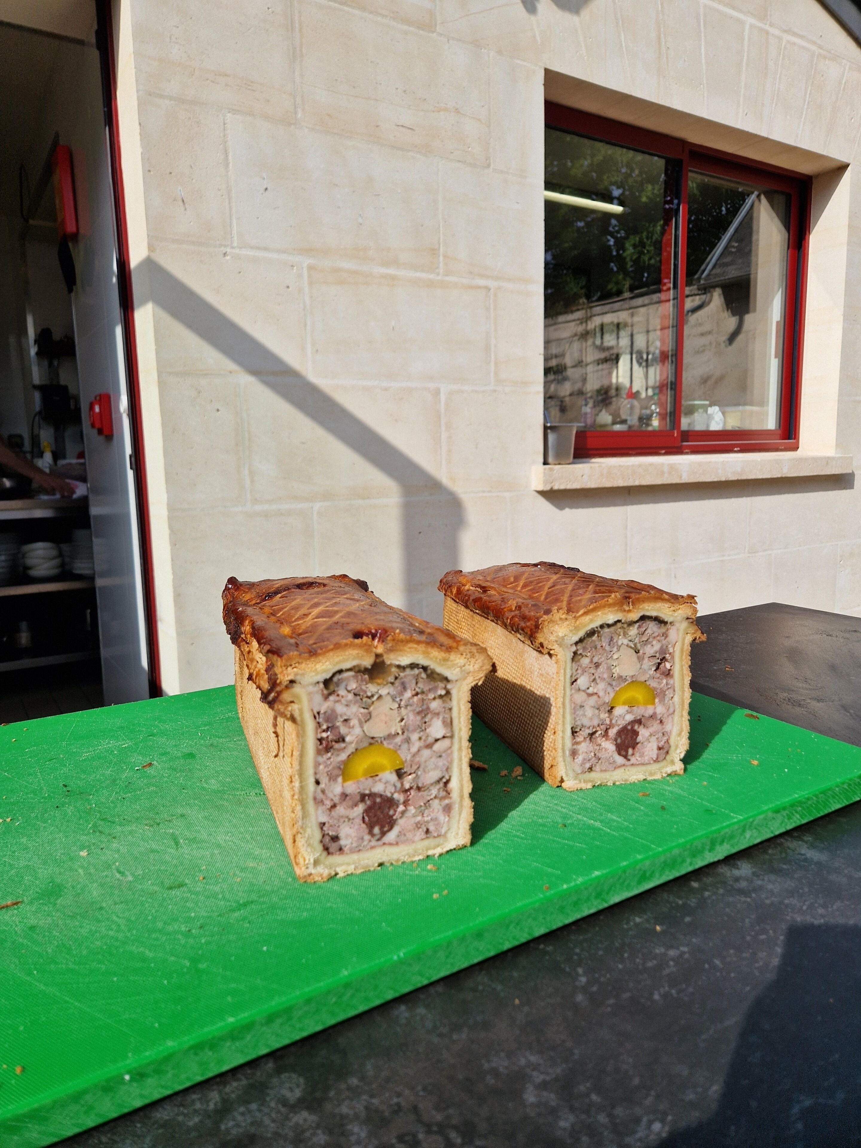 Two meat pies on a green cutting board in front of a building with a window.