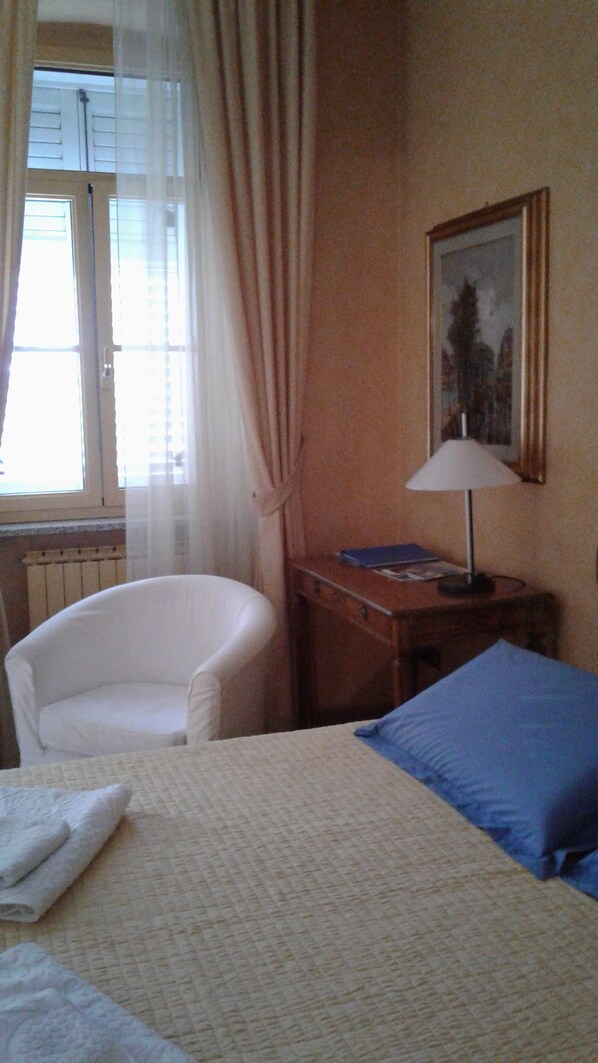 Une chambre d’hôtel avec un lit, une chaise, une table avec une lampe, un tableau au mur et une fenêtre avec des rideaux.