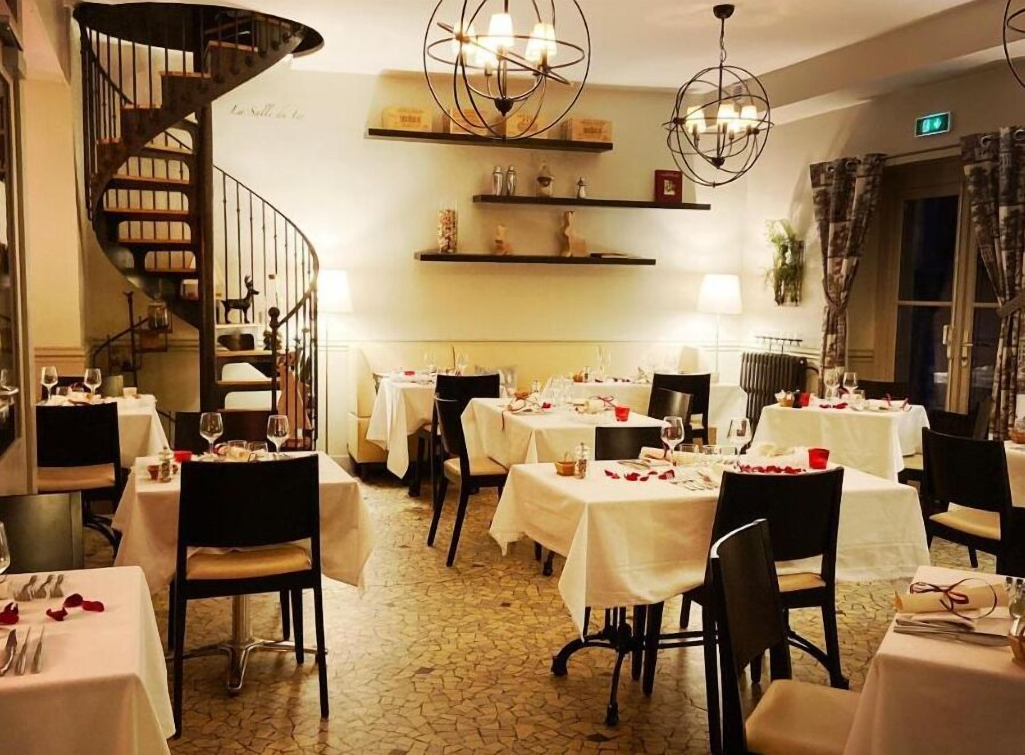 L’intérieur d’un restaurant, avec de nombreuses tables dressées pour le service, un escalier en colimaçon et des luminaires suspendus.