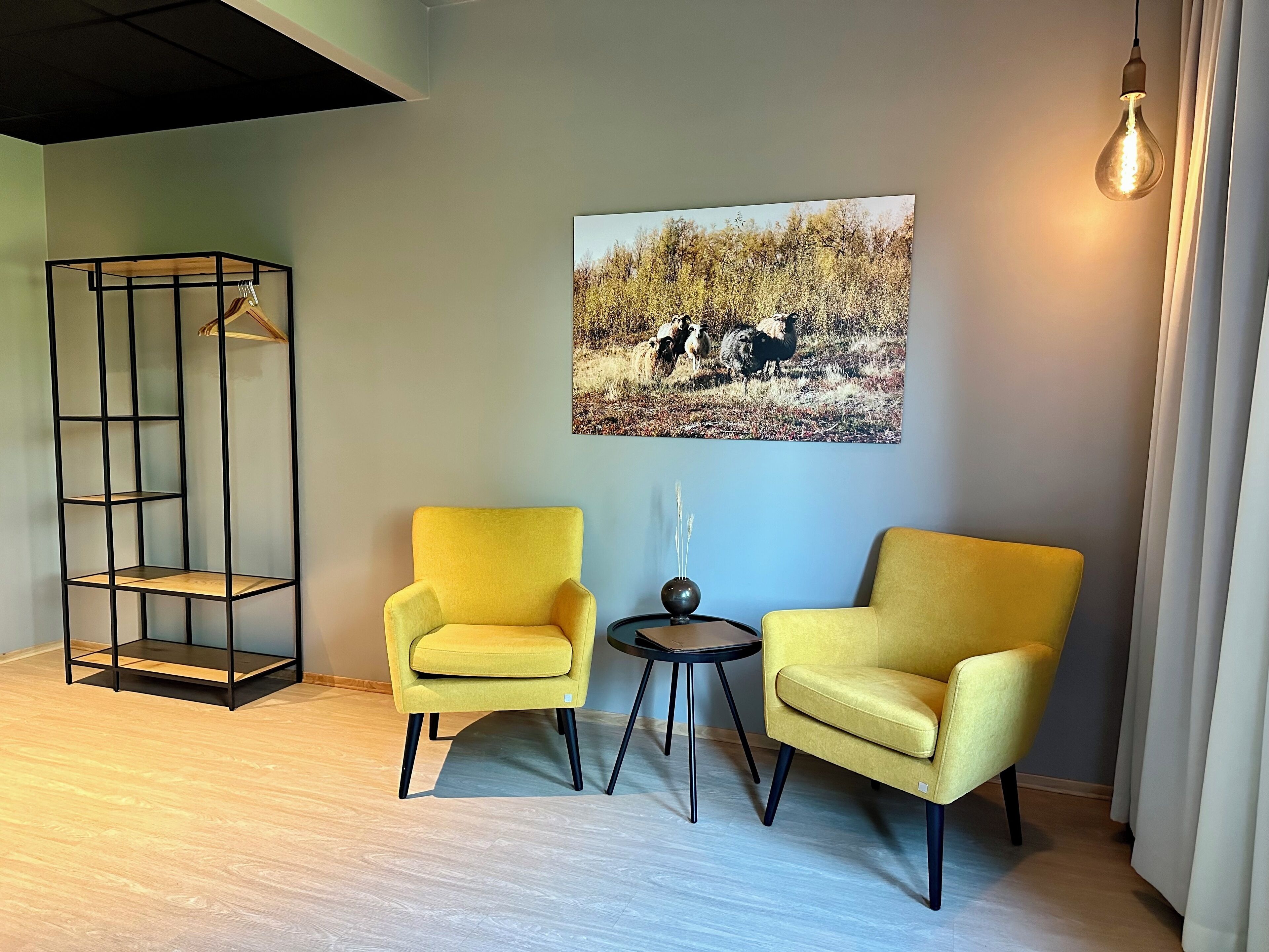 A room with two yellow armchairs, a black metal shelving unit, a round table with a vase, and a wall-mounted picture of sheep in a field.
