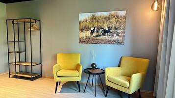A room with two yellow armchairs, a black metal shelving unit, a round table with a vase, and a wall-mounted picture of sheep in a field.