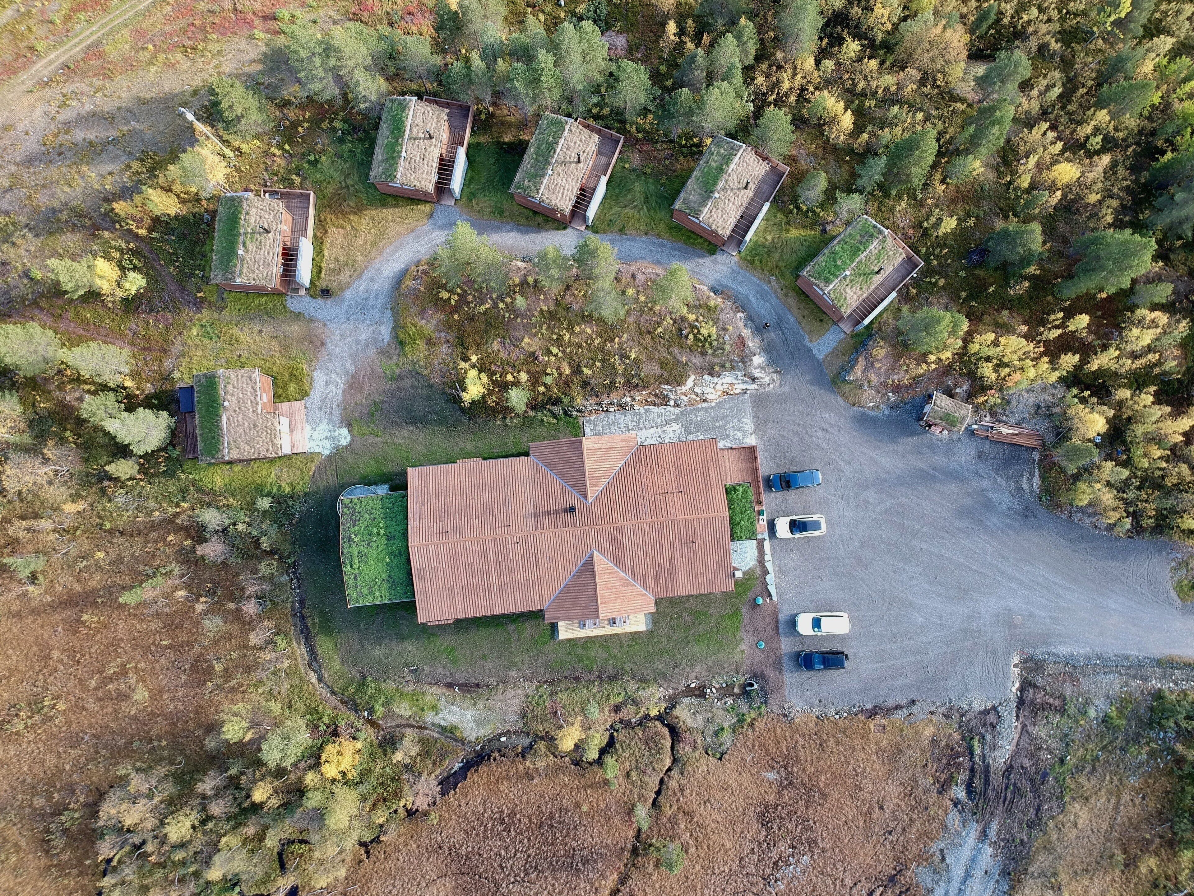 An aerial view of a property with multiple buildings, a large central building with a sloped roof, and a parking area with several cars.