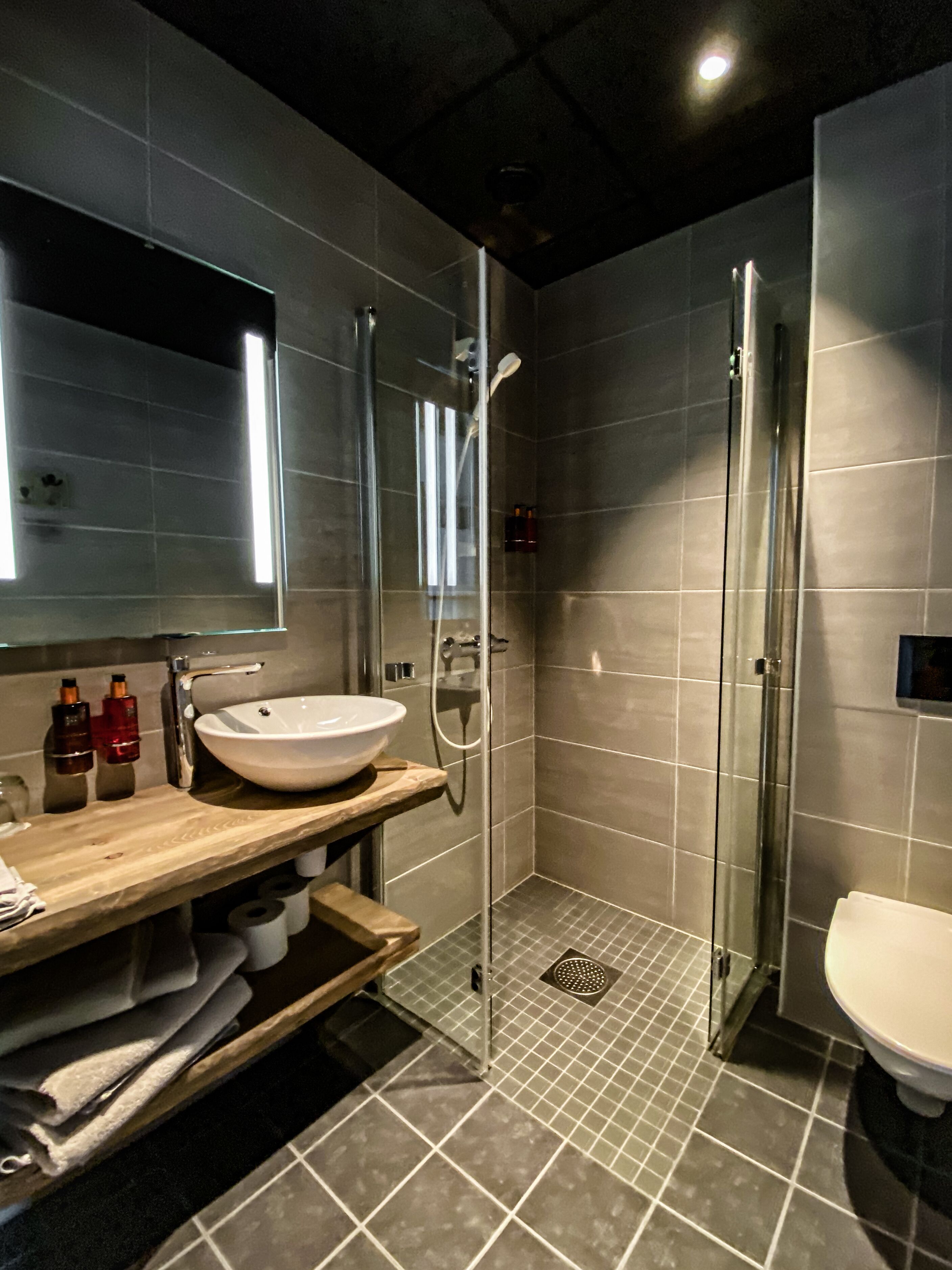 Un baño moderno con un espejo grande, un tocador de madera, un lavabo blanco, cabina de ducha de cristal y un inodoro.