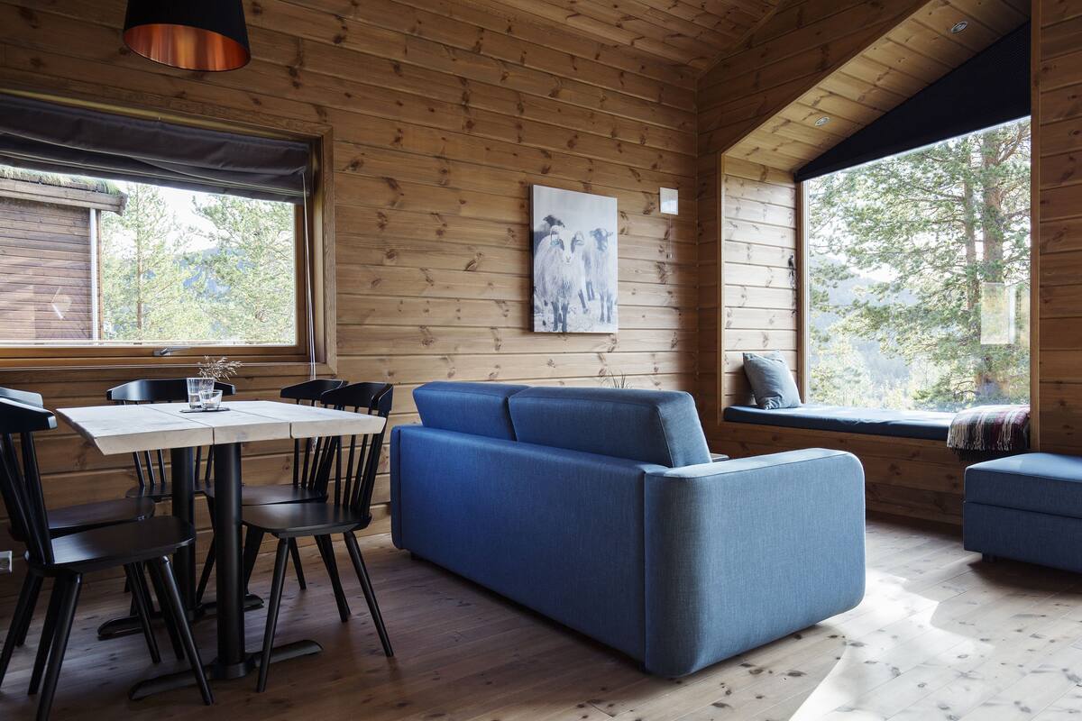 A wooden cabin interior with a dining table, chairs, a blue sofa, and a window with a view of trees.