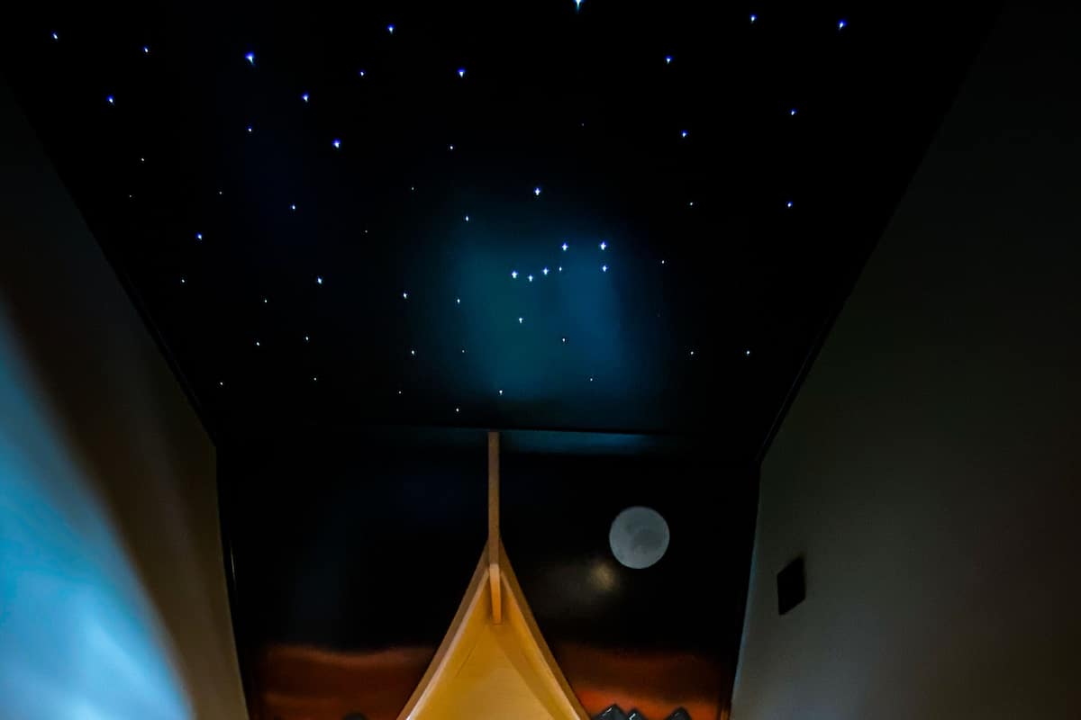 A room with a bed, a view of the night sky, and a moon.