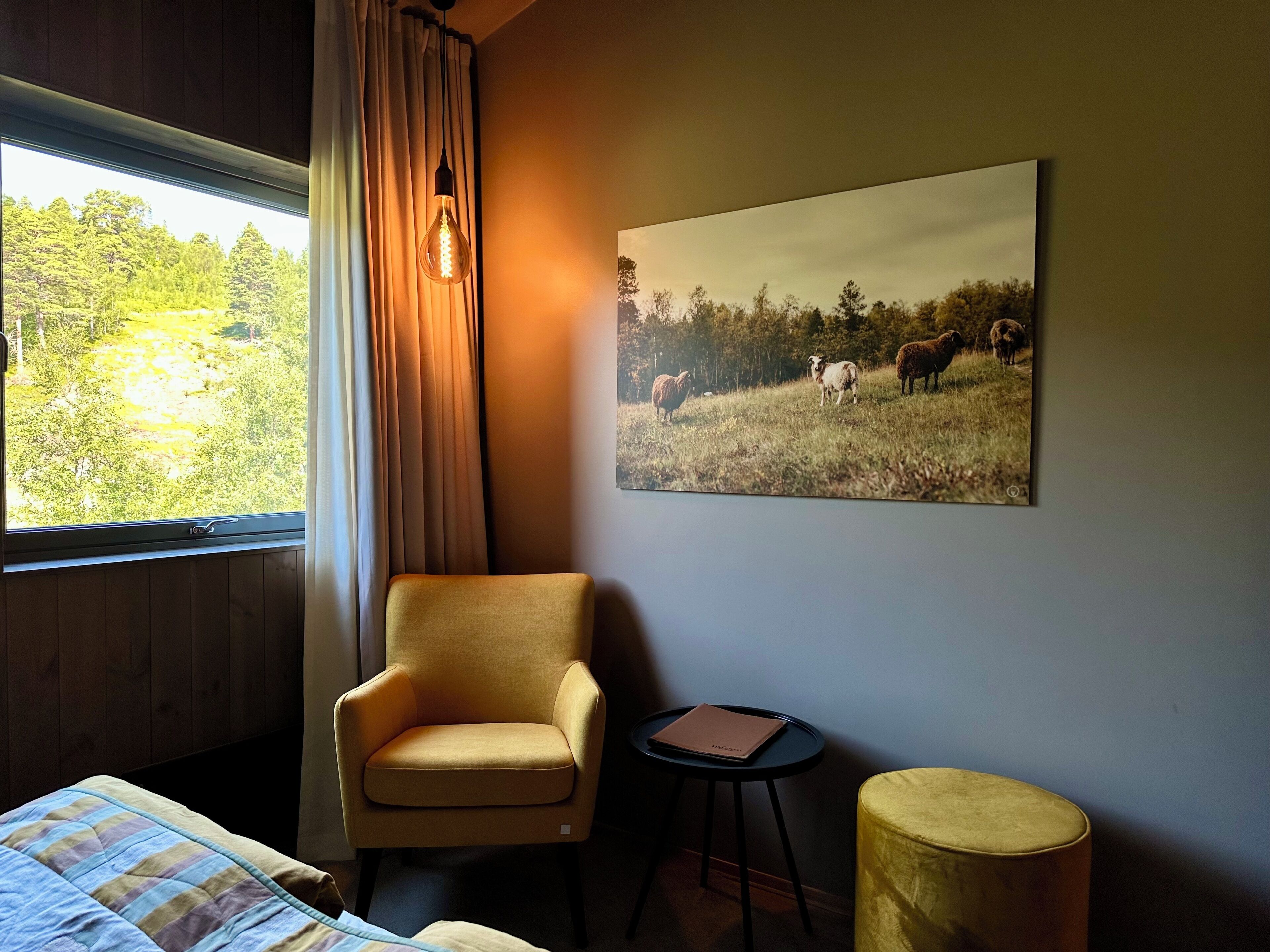 A room with a bed, a chair, a small table with a folder, a round yellow ottoman, a window with curtains, and a painting of sheep on a field.