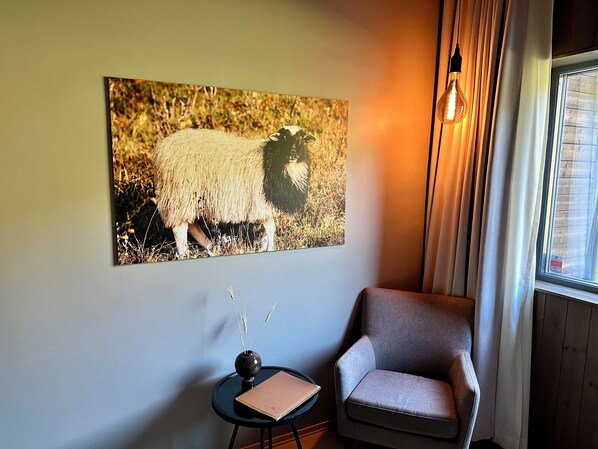 Una habitación con una obra de arte enmarcada de una oveja, una mesita redonda con un florero y una libreta, y un sillón gris.