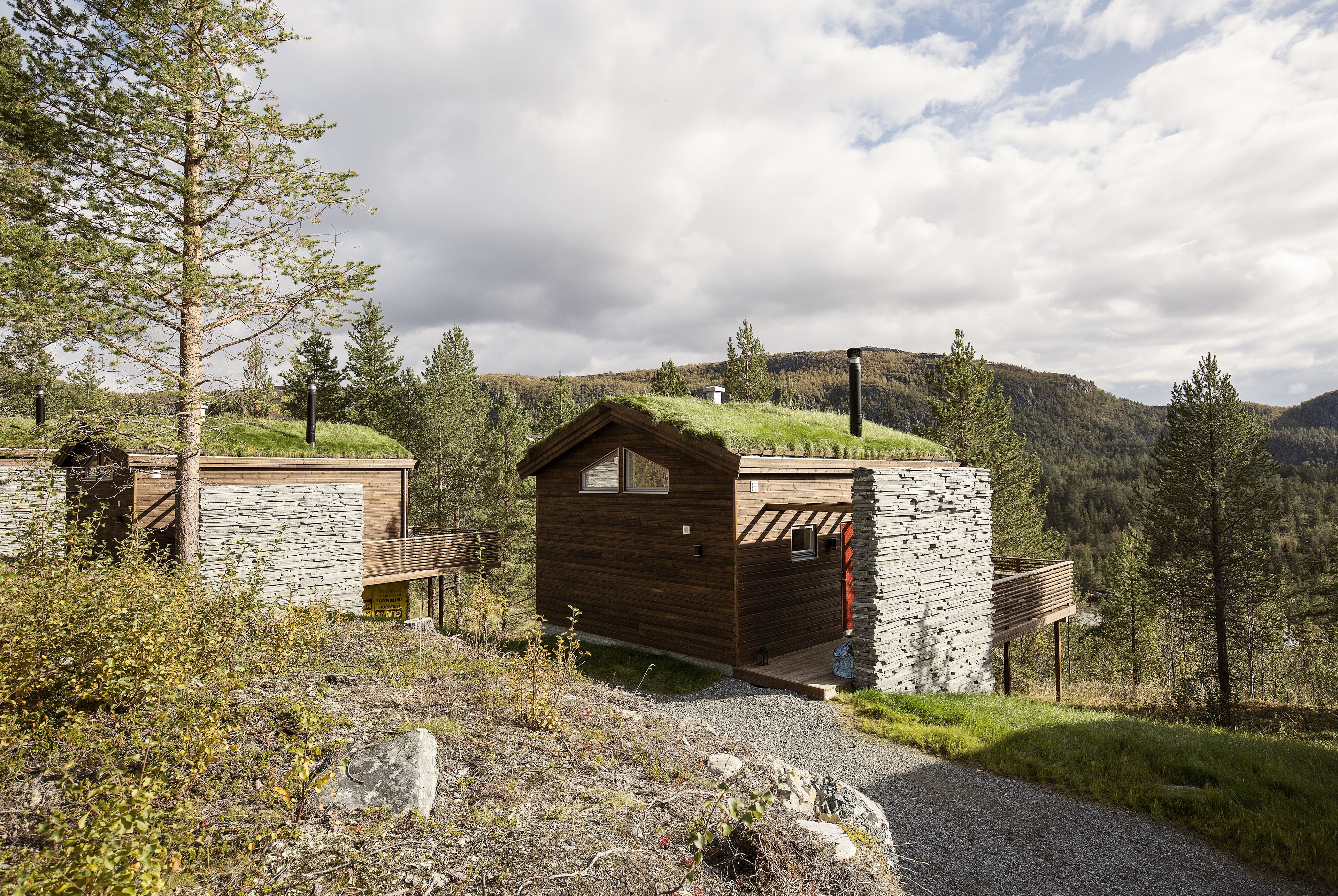 A rustic cabin with a stone exterior and wooden roof, surrounded by a natural landscape with trees and hills.