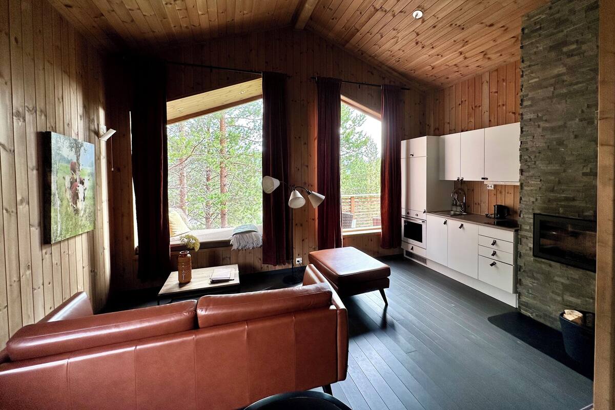 A cozy cabin interior with wooden walls, a stone fireplace, a dining area, and a kitchen.